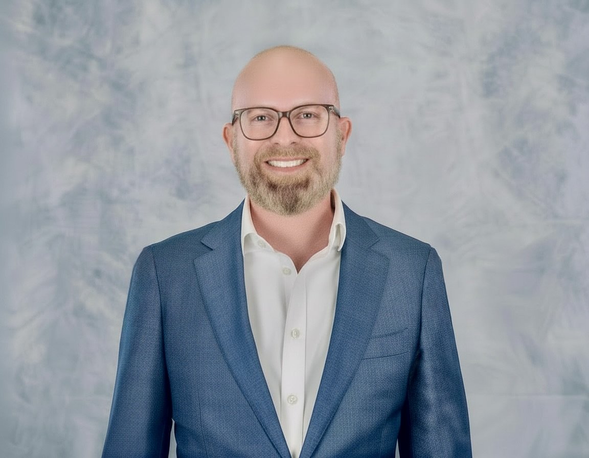Daniel Kaniewski is smiling and wearing a blue blazer and white shirt, standing against a light, cloudy background.
