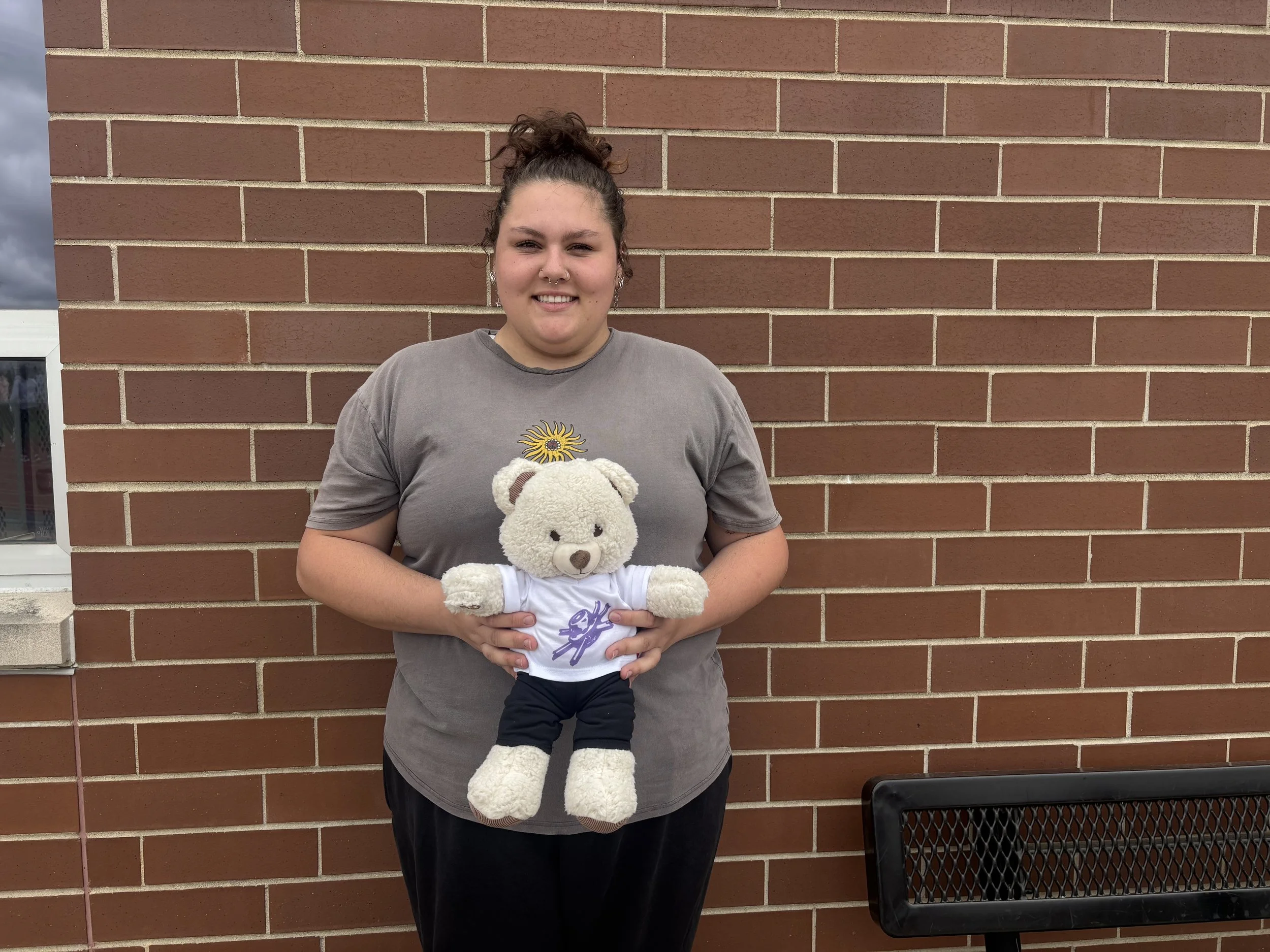 A woman with curly hair tied in a bun, smiling, holding a plush teddy bear dressed in a sports uniform, standing against a brick wall.