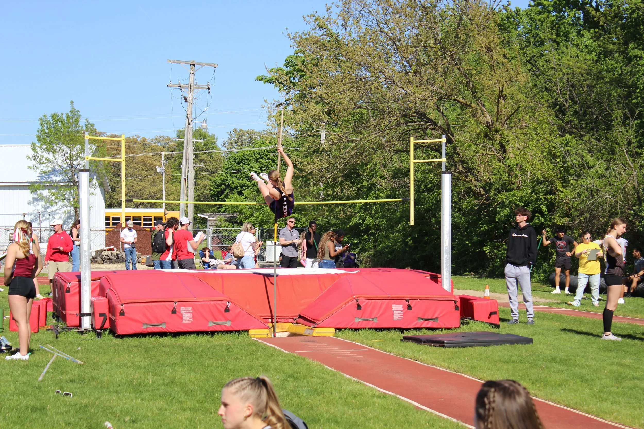 A female pole vaulter in mid-air during her jump at an outdoor track meet, with spectators and other athletes watching nearby.