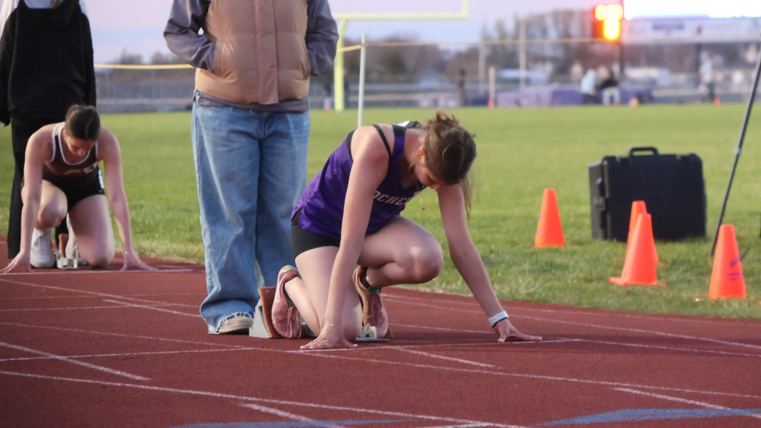 A female athlete in a purple track uniform kneels at the starting line on a red running track, preparing for a race. She is in a crouched position, with one hand on the ground and wearing running shoes. An official or coach, wearing jeans and a brown