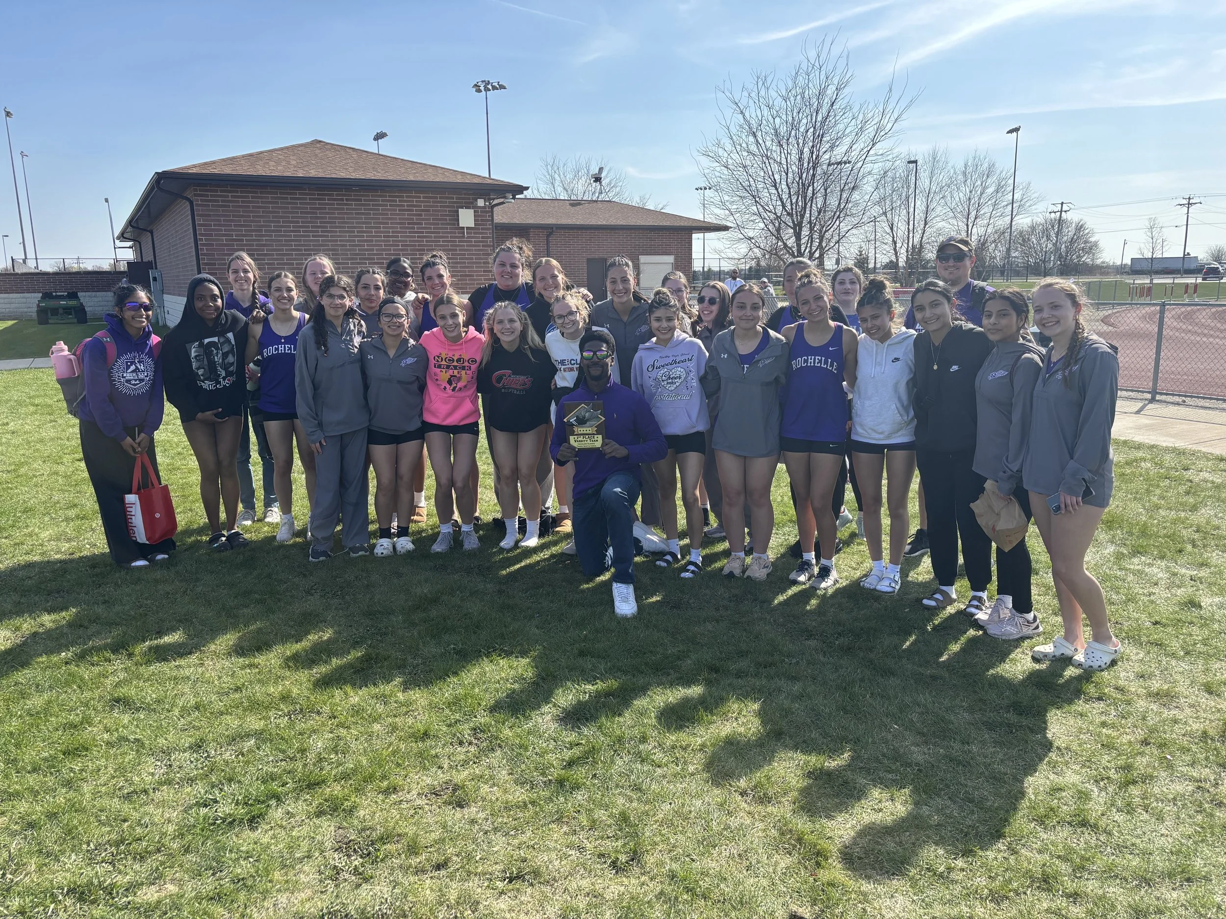 Group of young female athletes in athletic gear and hoodies, standing on grass field at track and field stadium, some holding a plaque, with a coach or team leader kneeling in front, on a sunny day.