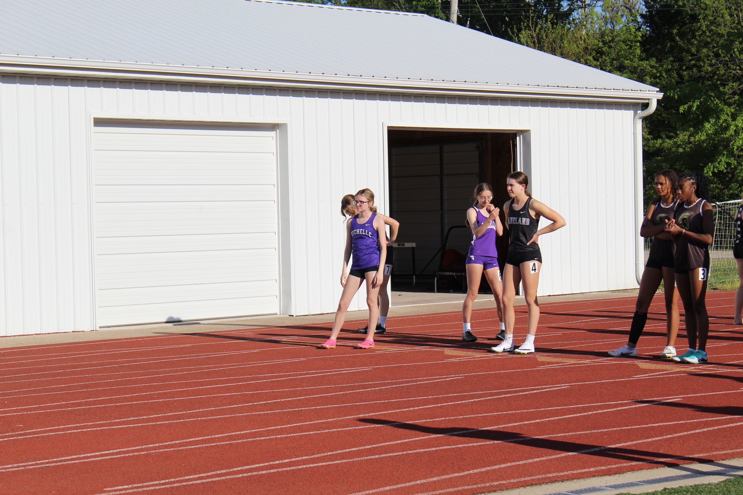 Young female athletes preparing for a race at a track and field event, standing in starting positions on a red running track, with a white building and green trees in the background.