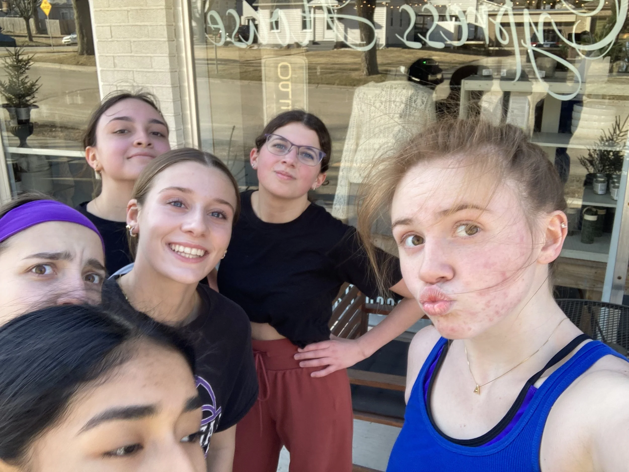 Group of six young women taking a selfie outside a shop window on a sunny day, with a street and trees visible in the background.