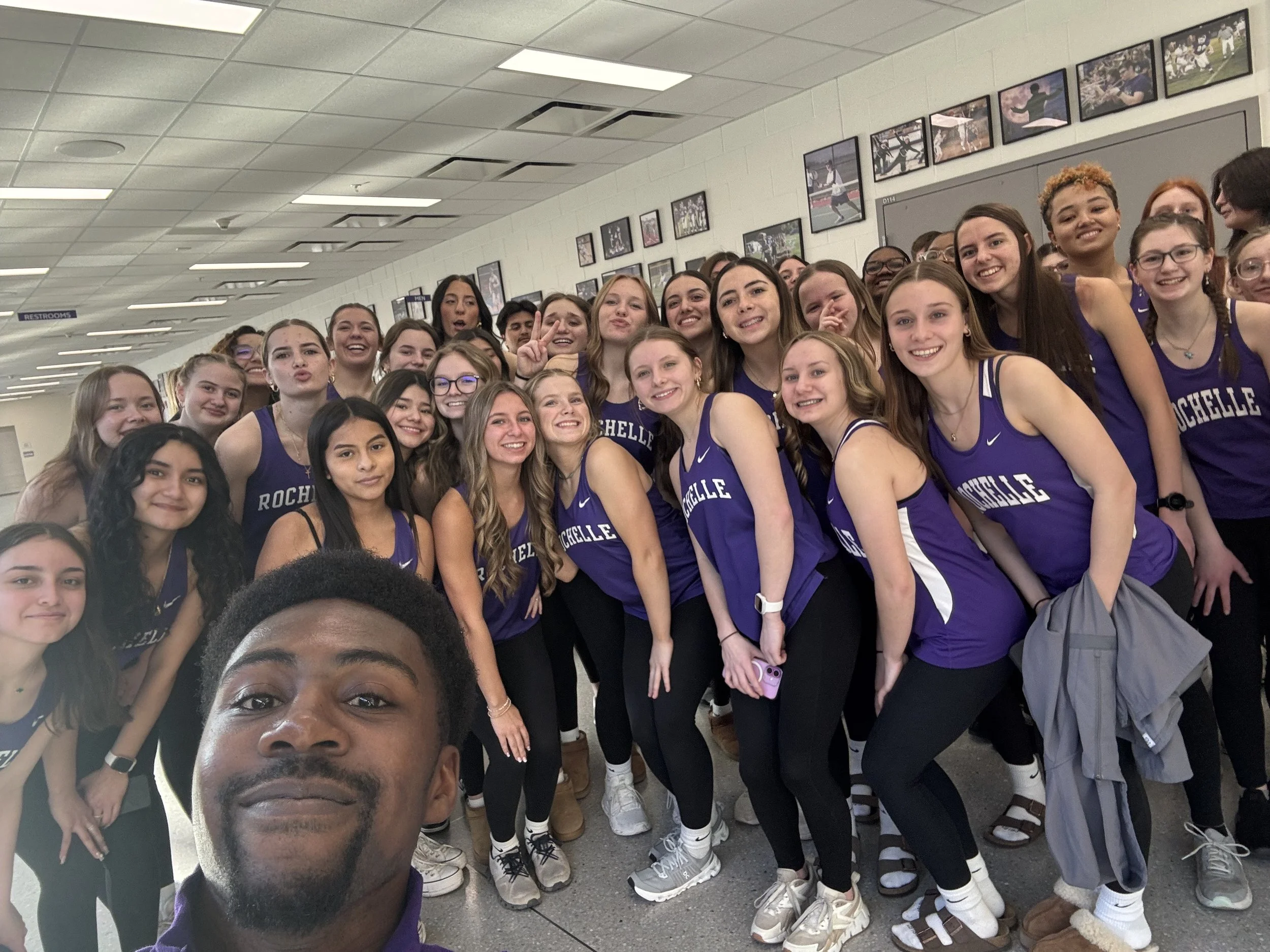 A group of young women wearing matching purple basketball jerseys with 'ROCHELLE' written on them, posed for a selfie in a hallway decorated with framed photos.