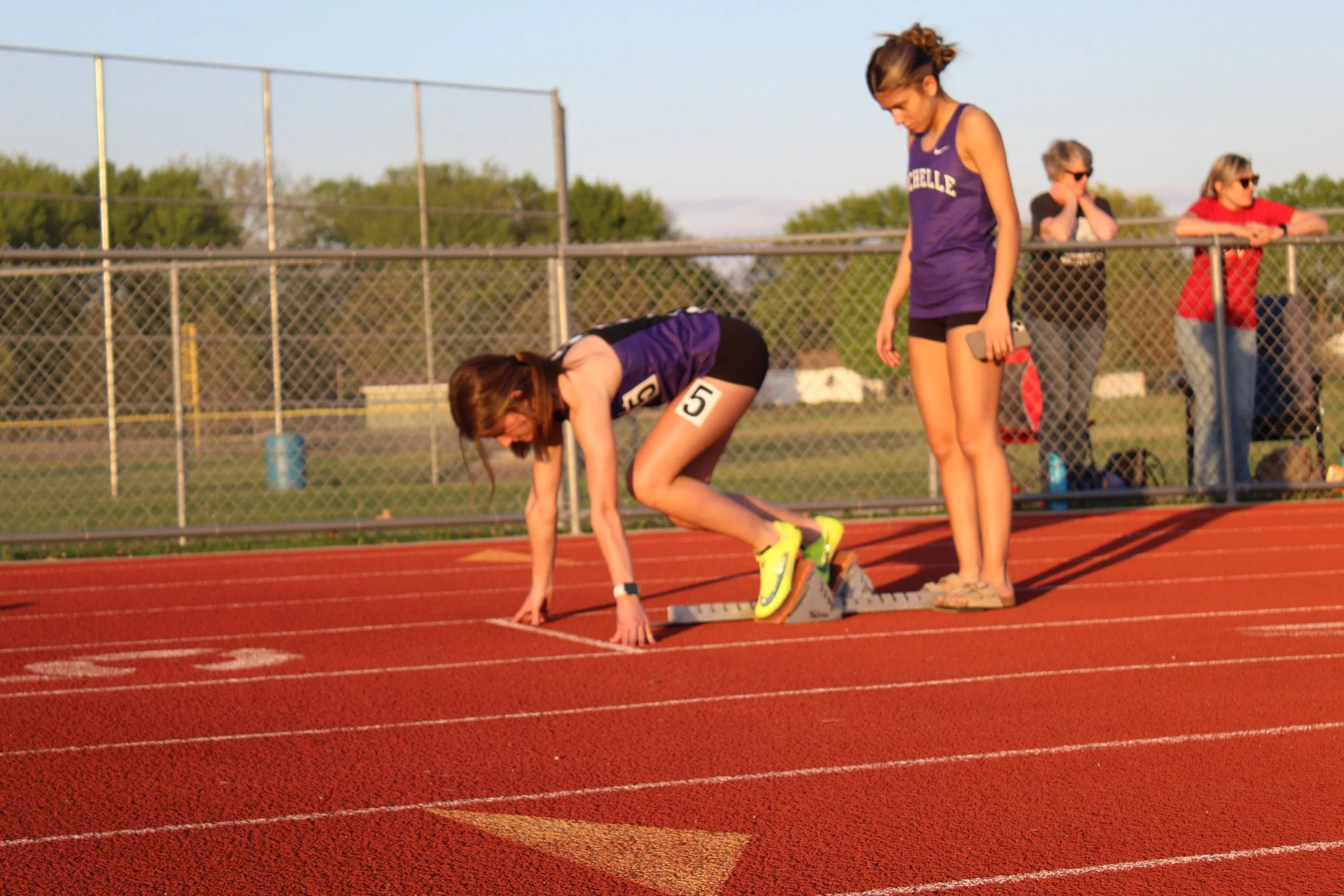 A female runner in purple athletic attire crouches at the starting line on a red track, preparing to race, while another woman in purple stands behind her holding a smartphone. Three spectators watch from behind a chain-link fence in the background d