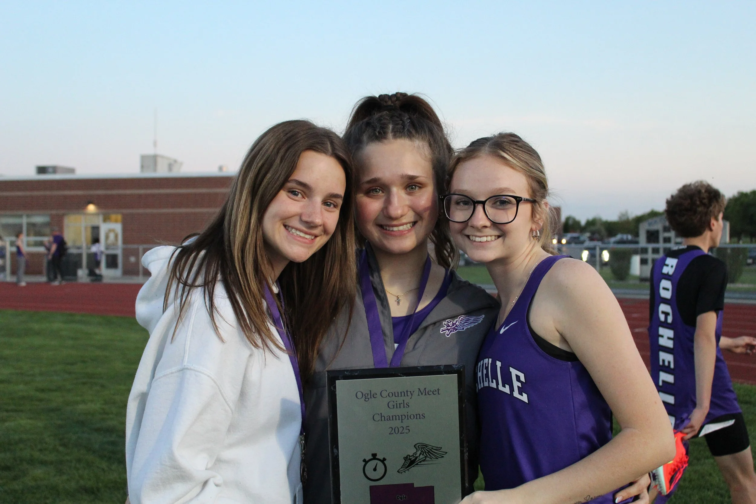 Three smiling teenage girls, two wearing glasses, celebrating their victory holding a plaque that reads 'Ogle County Meet Girls Champions 2025' during a sports event on a track field at dusk.