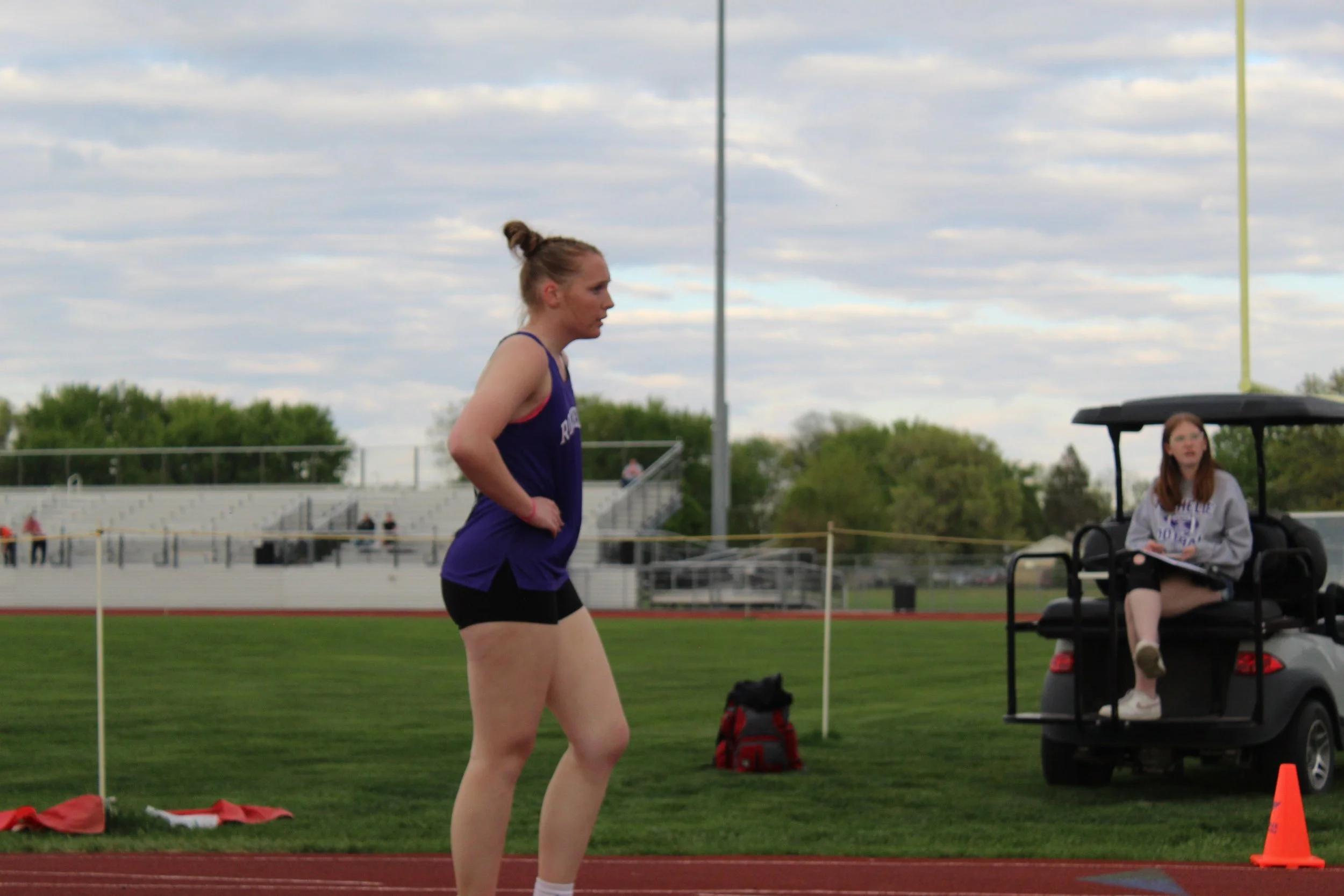 A female athlete stands on a running track with hands on her hips, wearing a purple sports top and black shorts. A woman sits on a golf cart nearby with a clipboard. The background features a bleacher seating area, green field, trees, and a cloudy sk