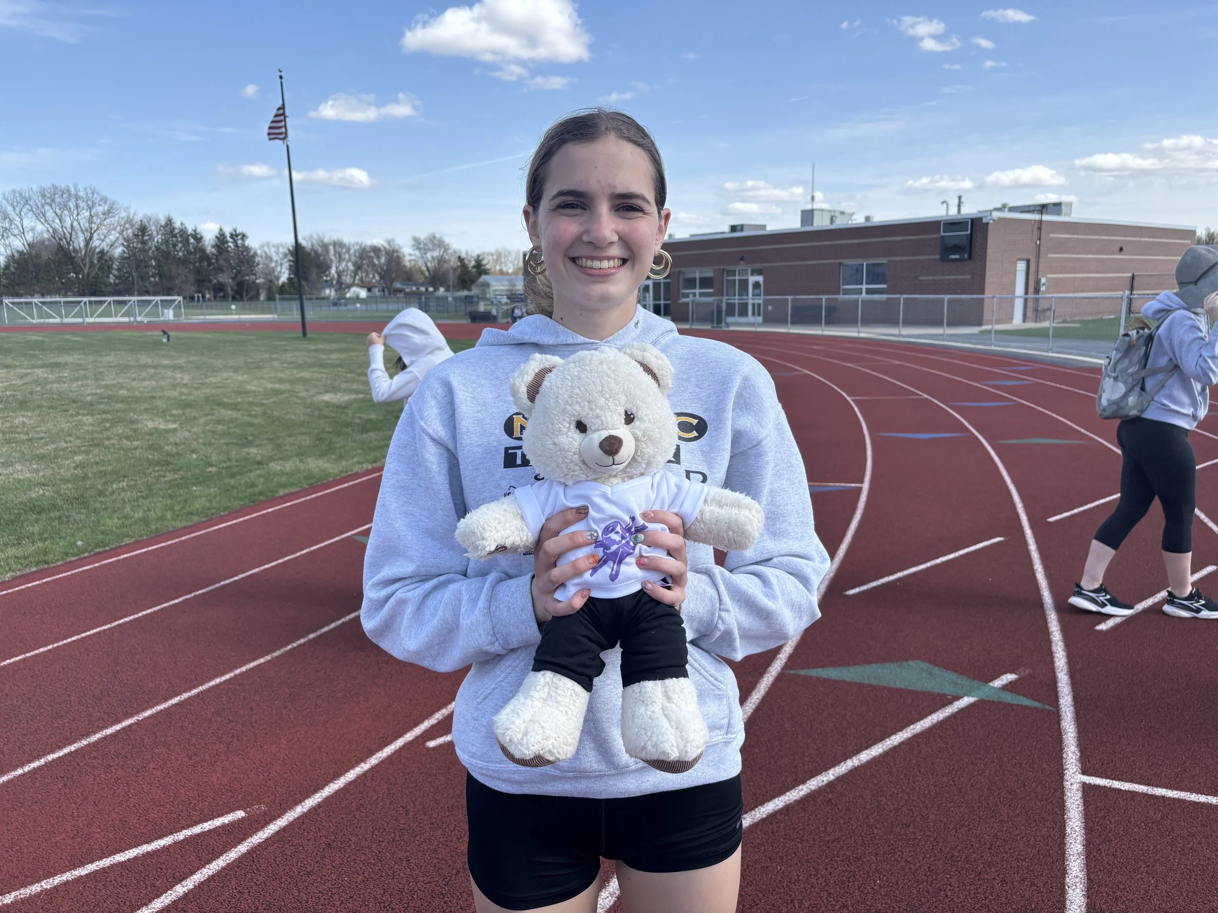 A young woman holding a teddy bear on a running track at a school stadium, with other students in the background, blue sky, and some buildings.
