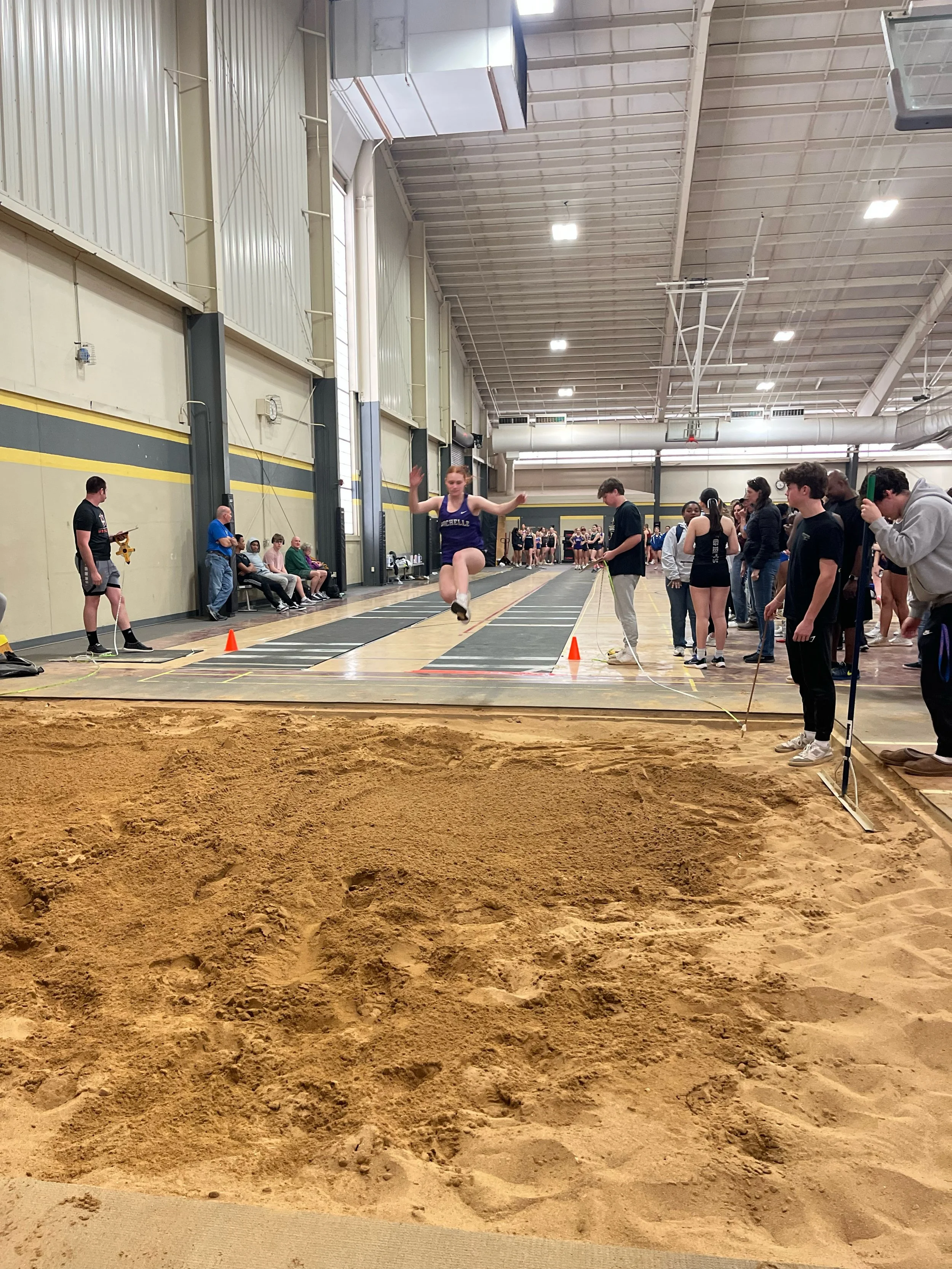 Indoor long jump competition with a female athlete mid-air landing in the sandpit, surrounded by spectators and officials.