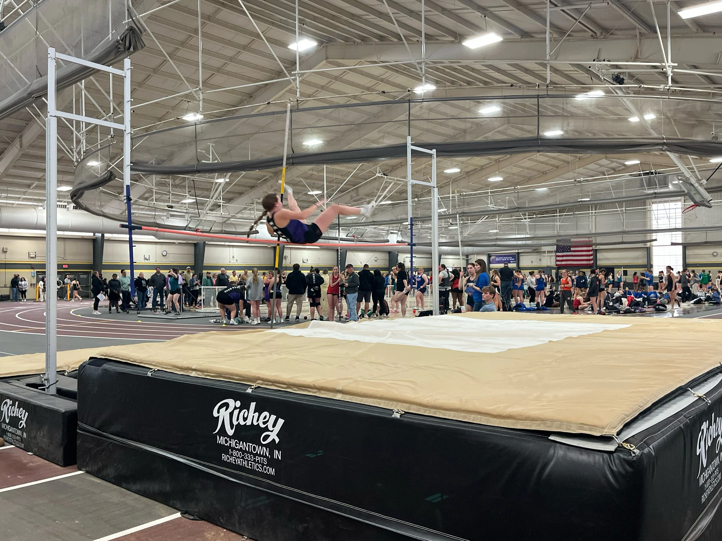 Indoor track and field event with athletes, spectators, and a high jump area featuring a female athlete in mid-air during a high jump attempt.