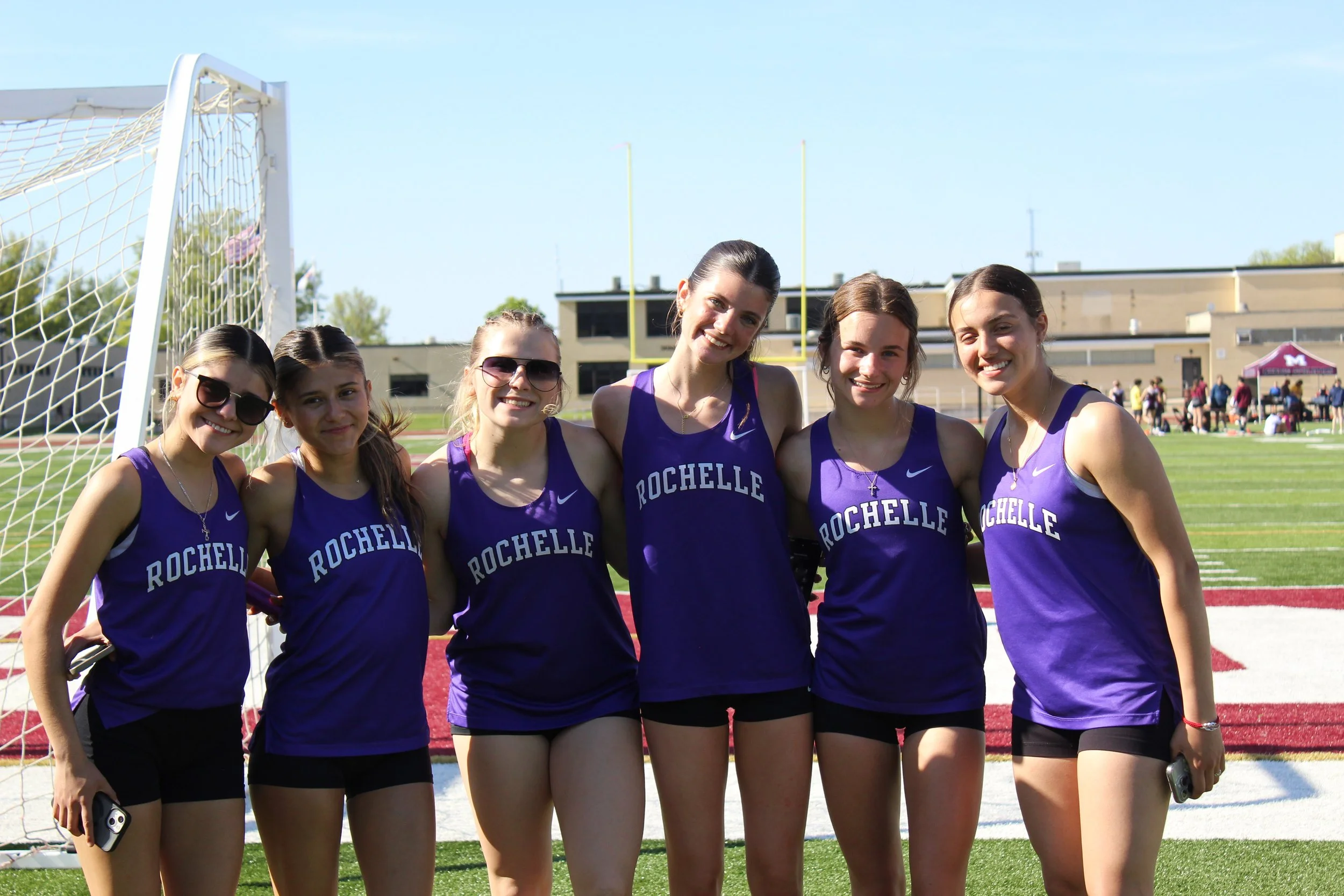 Six female track athletes in purple uniforms labeled 'Rochelle' standing on a track in front of a goalpost, smiling at the camera.