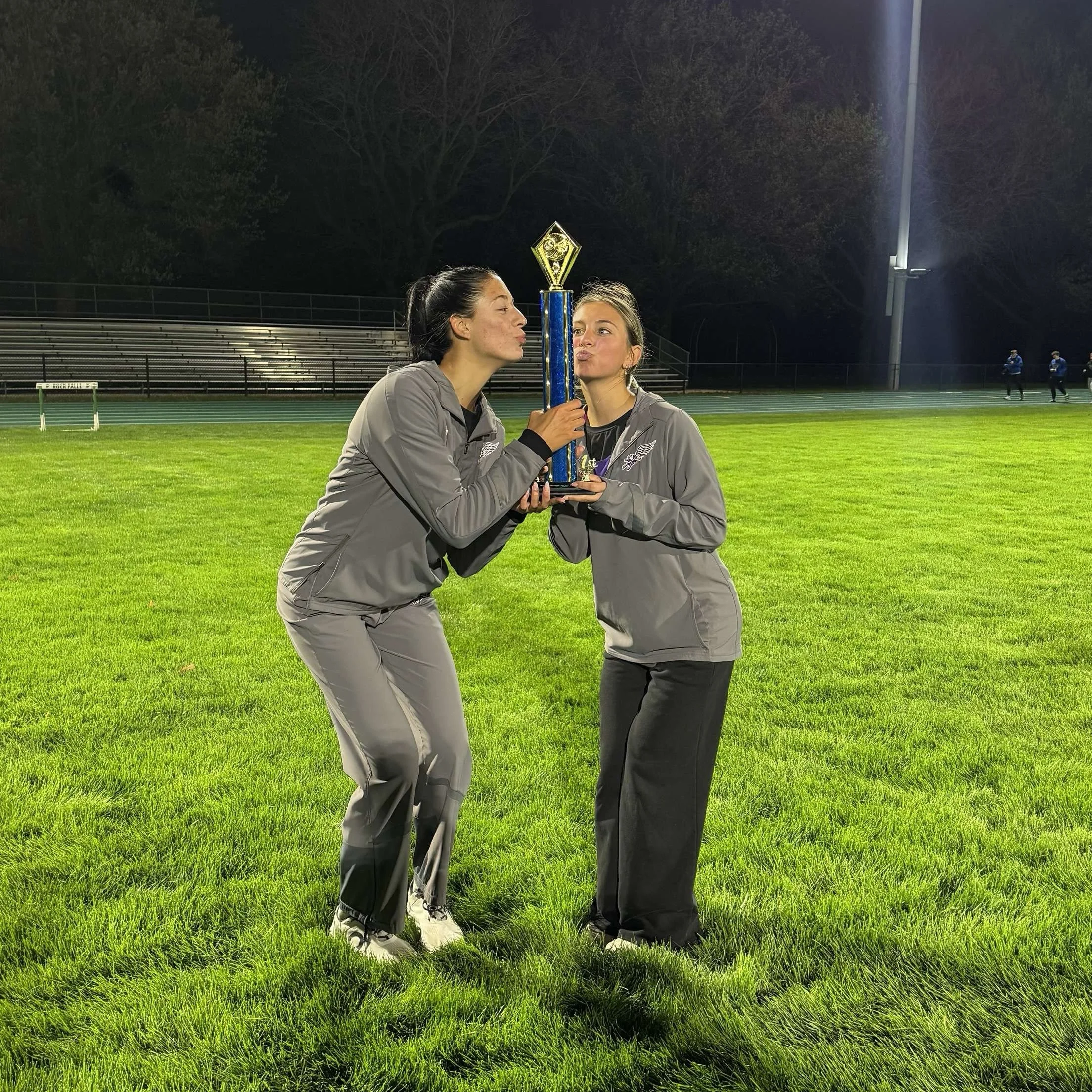 Two female athletes wearing gray sports jackets and black pants holding a tall trophy together on a football field at night.