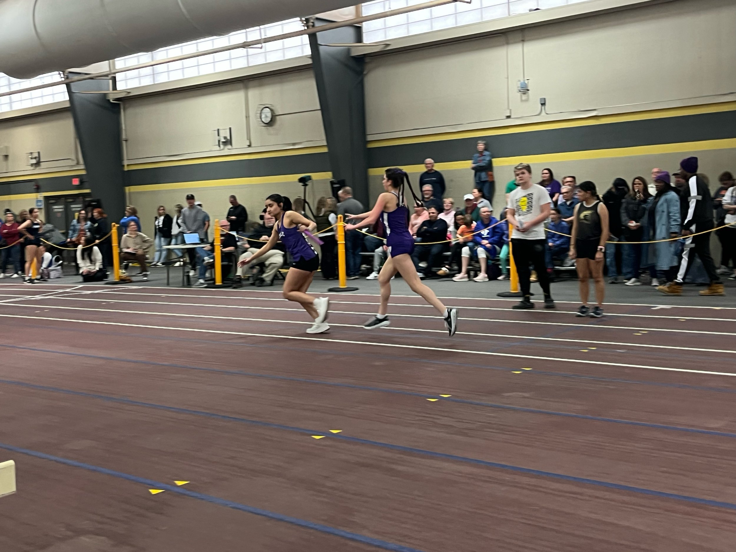 Two female runners in purple uniforms competing in an indoor track race, with a crowd of spectators and officials watching from the sidelines.