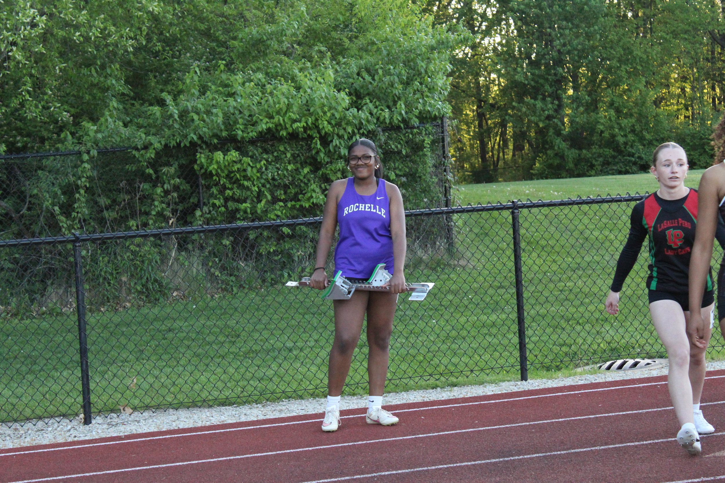 A young female athlete standing on a track holding her starting blocks, wearing a purple Rochelle sports uniform, smiling with glasses, near a chain-link fence and green trees in the background.