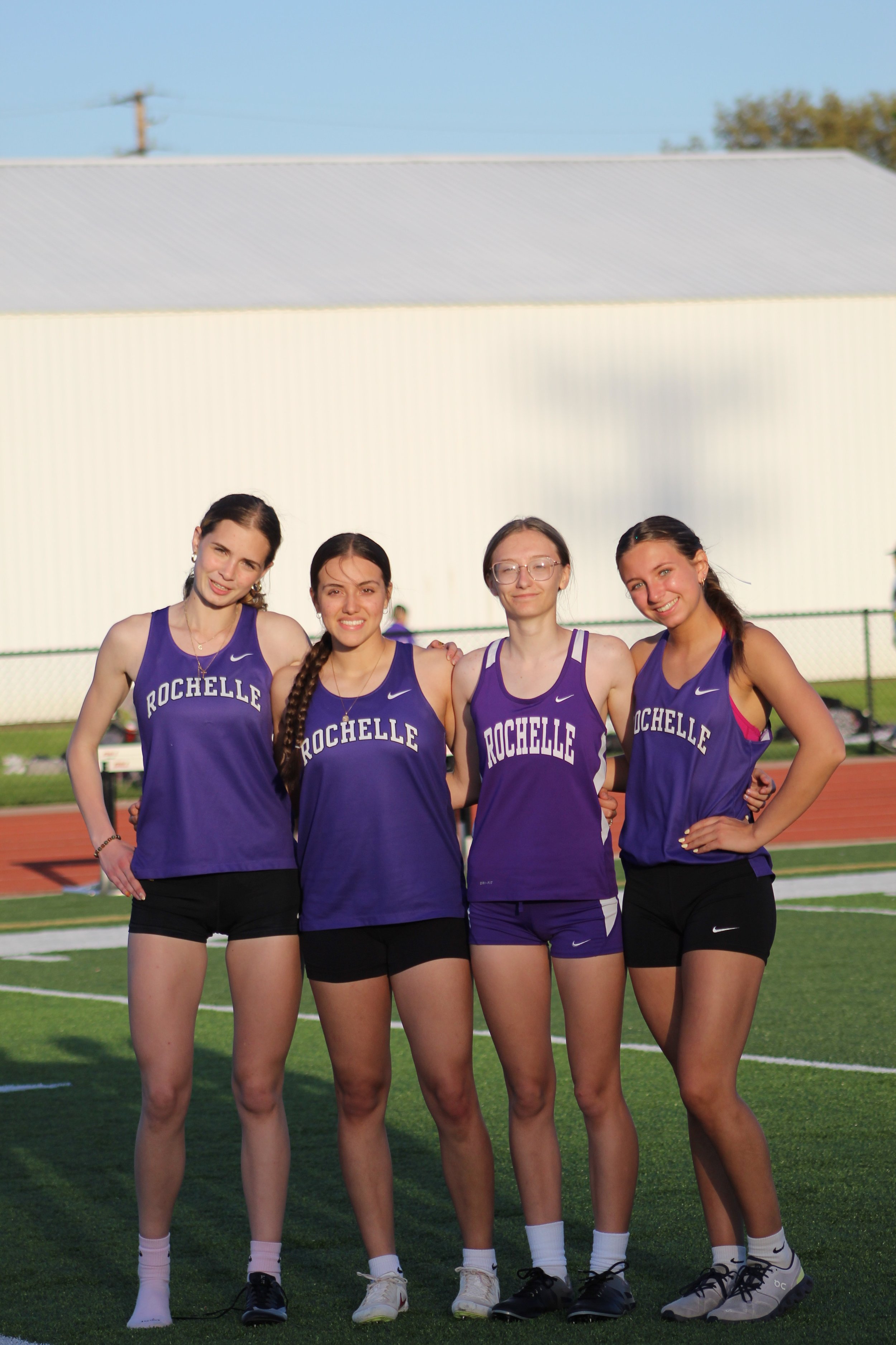 Four young female track athletes standing side by side outdoors, wearing purple Rochelle team uniforms, on a running track during daylight.