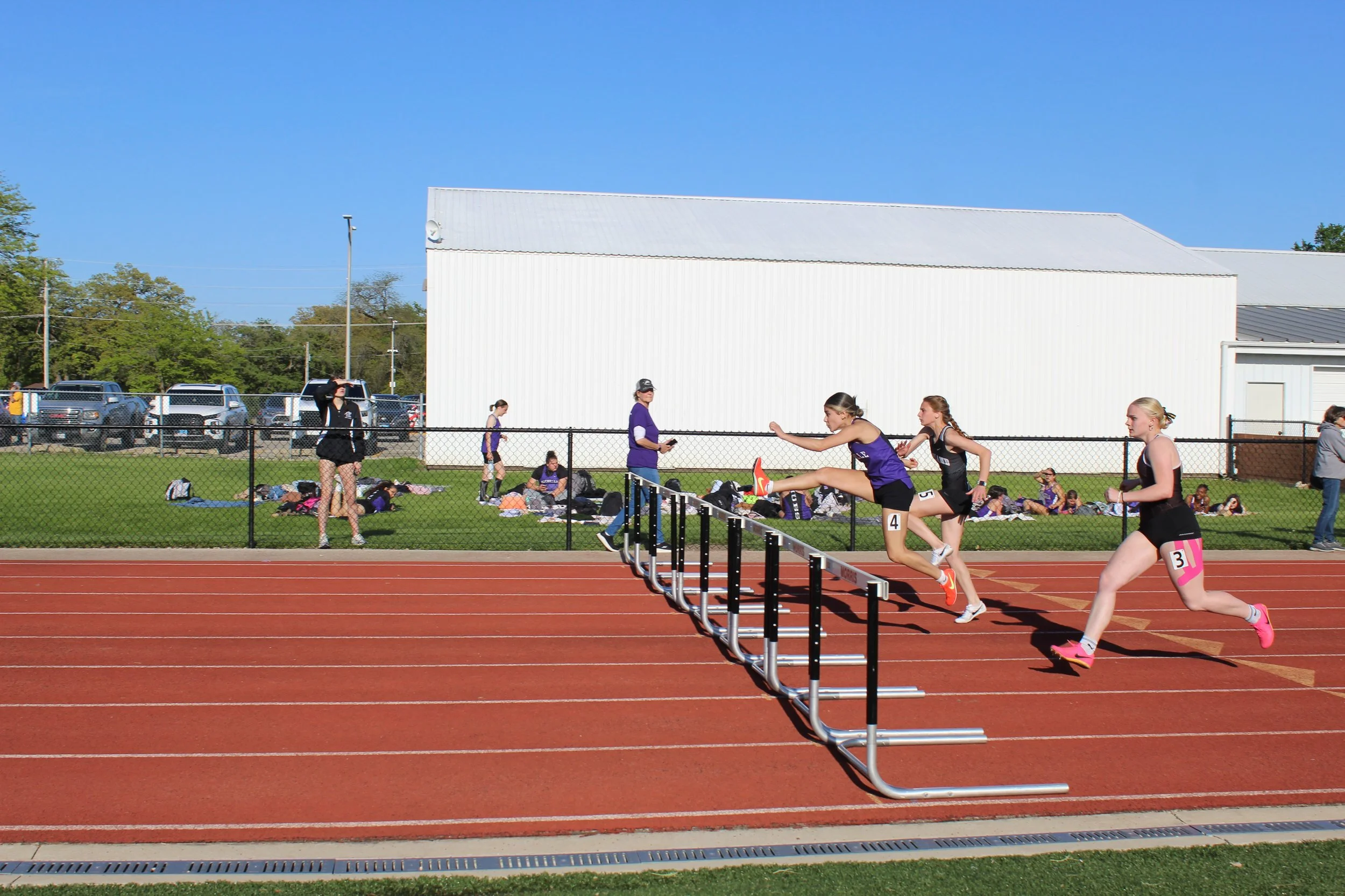 Young female athletes participating in a hurdle race on a red track at an outdoor sports facility. Some athletes are in mid-air over hurdles, others are running. Background includes a grassy area with spectators and coaches, a chain-link fence, parke