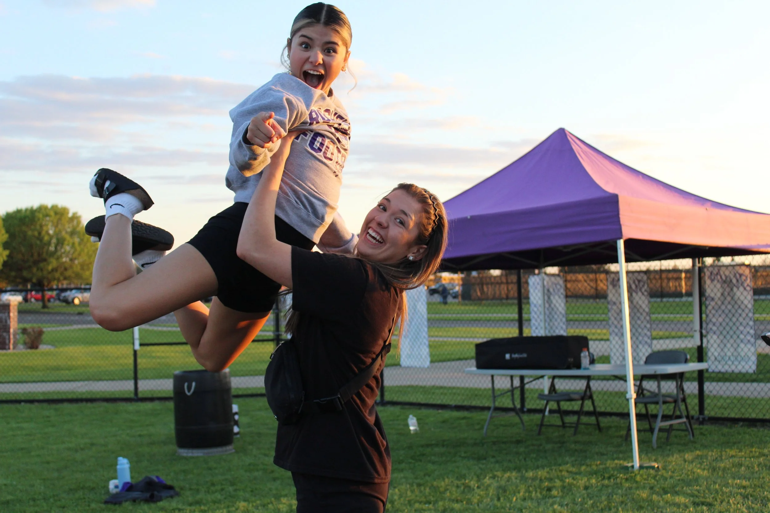 A woman cheerfully lifting a young girl in the air outdoors at sunset, with a purple tent and a fence in the background.
