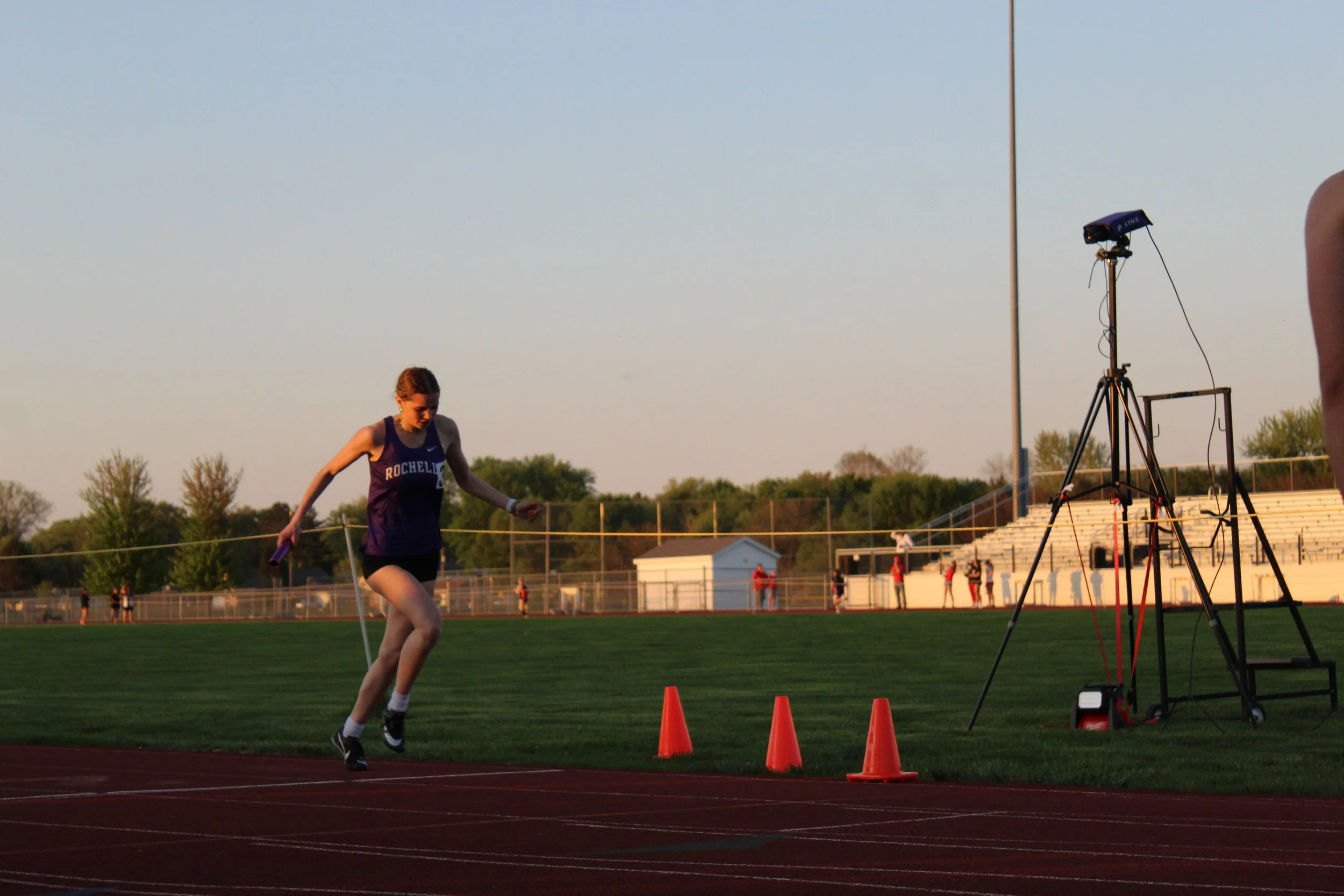 A female athlete runs on a track during a relay race, holding a purple baton. There are orange cones on the track and a timing camera on a tripod nearby. Spectators and other athletes are visible in the background as the sun sets.