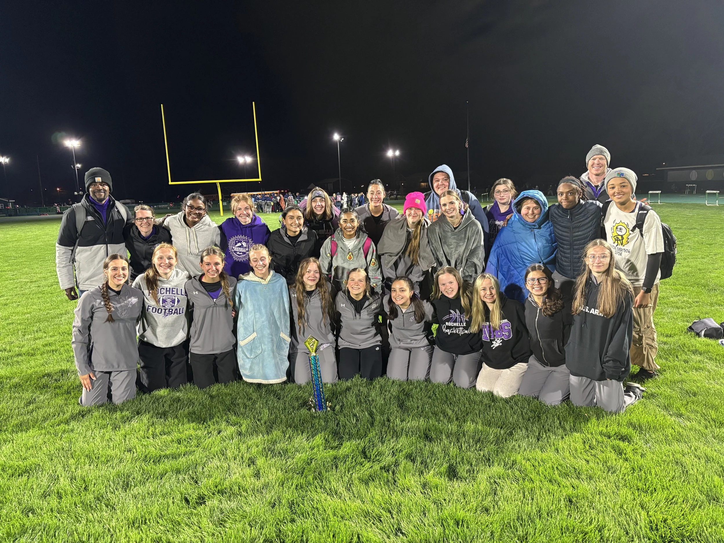 Group of young female athletes and coaches posing for a team photo on a football field at night, with a trophy placed in front of them. The group is dressed in sportswear, some wearing hoodies with team logos, and are smiling happily.