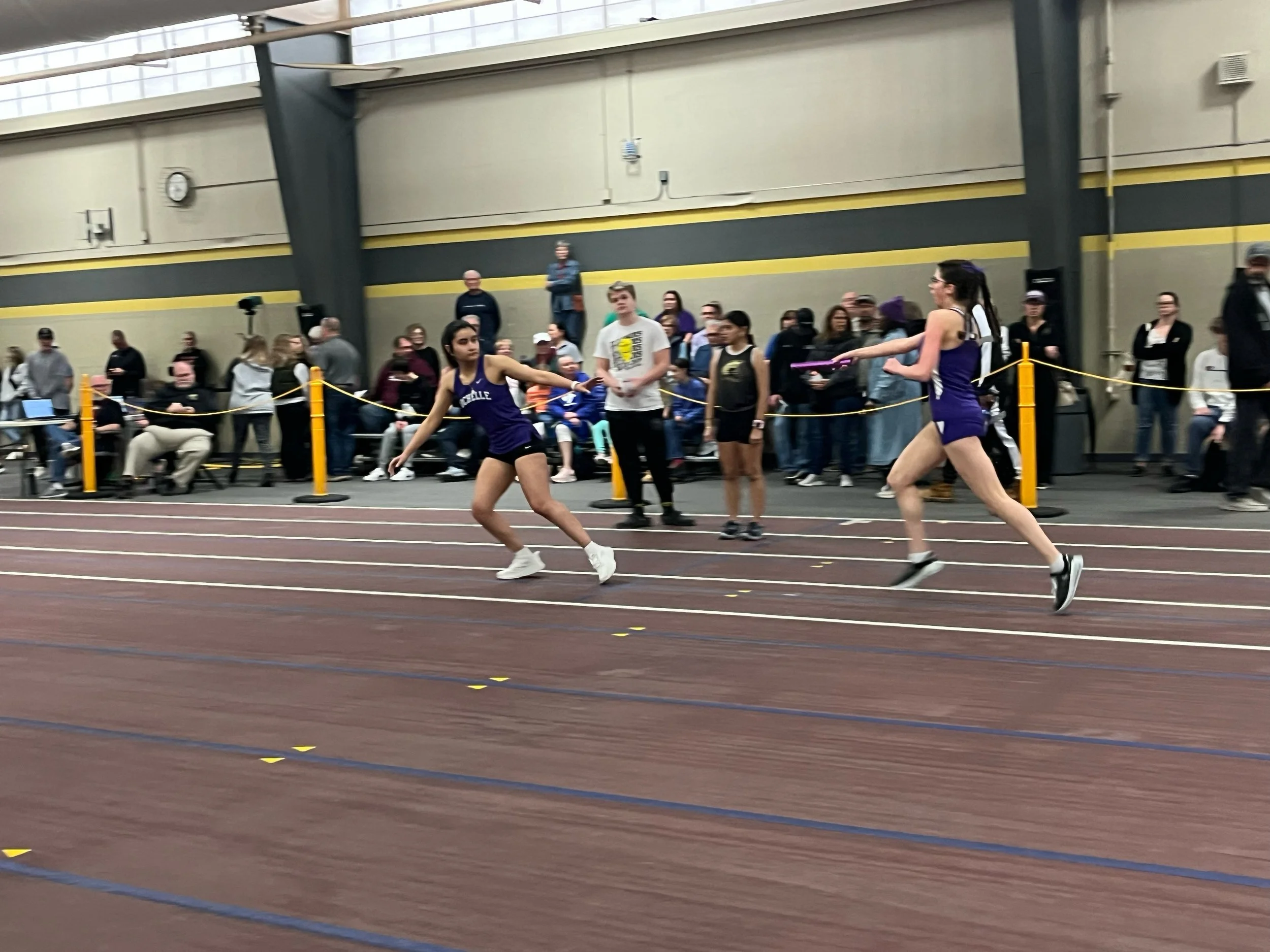 Two female athletes racing on an indoor track with an audience watching in the background.