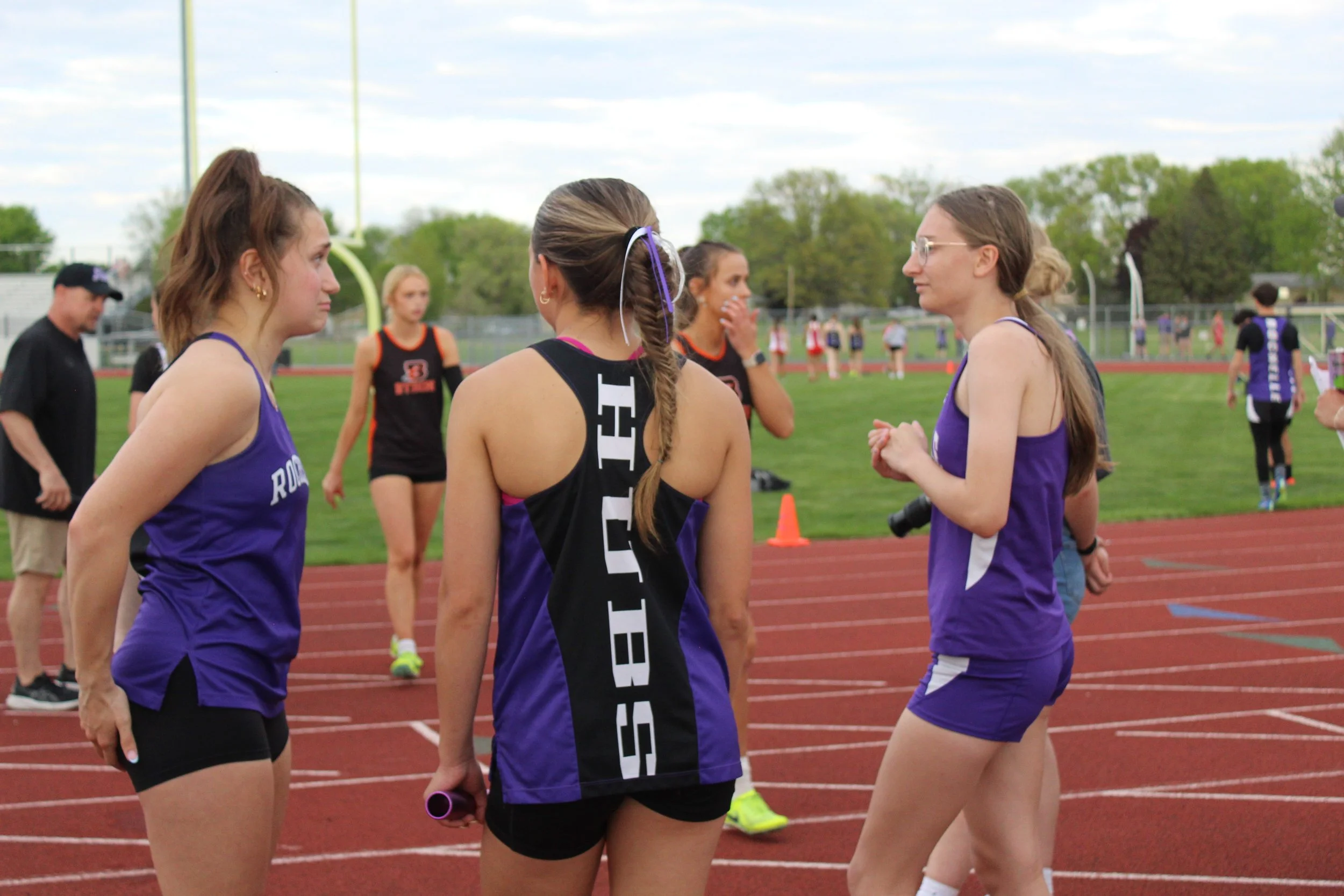 Track and field athletes in purple and black uniforms talking on a red running track during a meet, with green field and trees in the background.