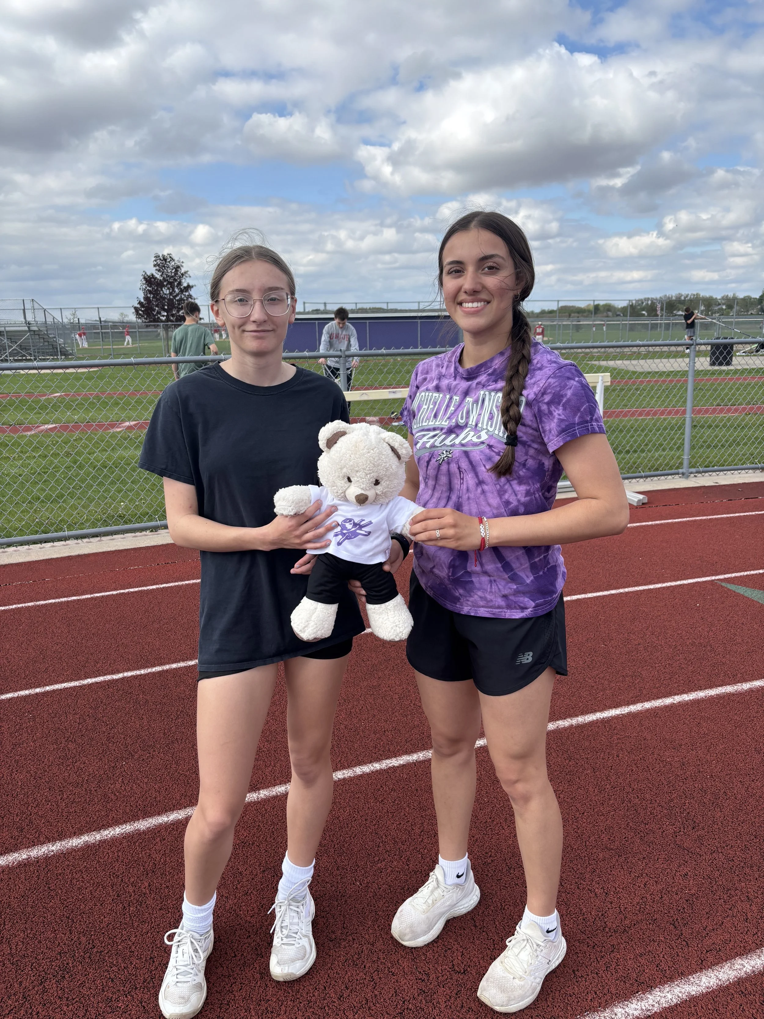 Two young women standing on a track field, one holding a teddy bear, with a chain-link fence and other athletes in the background under a partly cloudy sky.