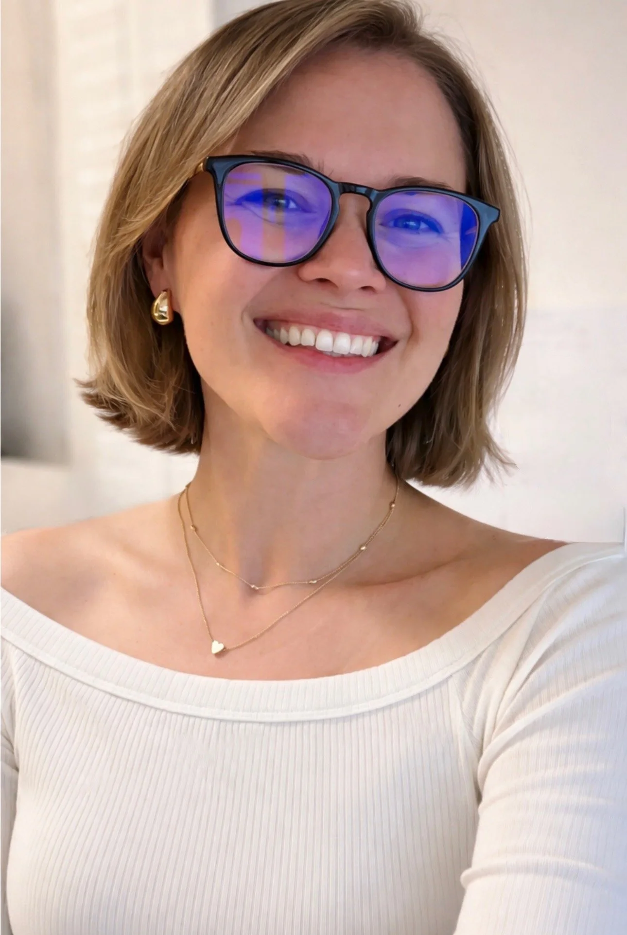A woman with short blonde hair, wearing glasses with blue light filters, gold earrings, and layered gold necklaces, smiling in front of a light-colored background.
