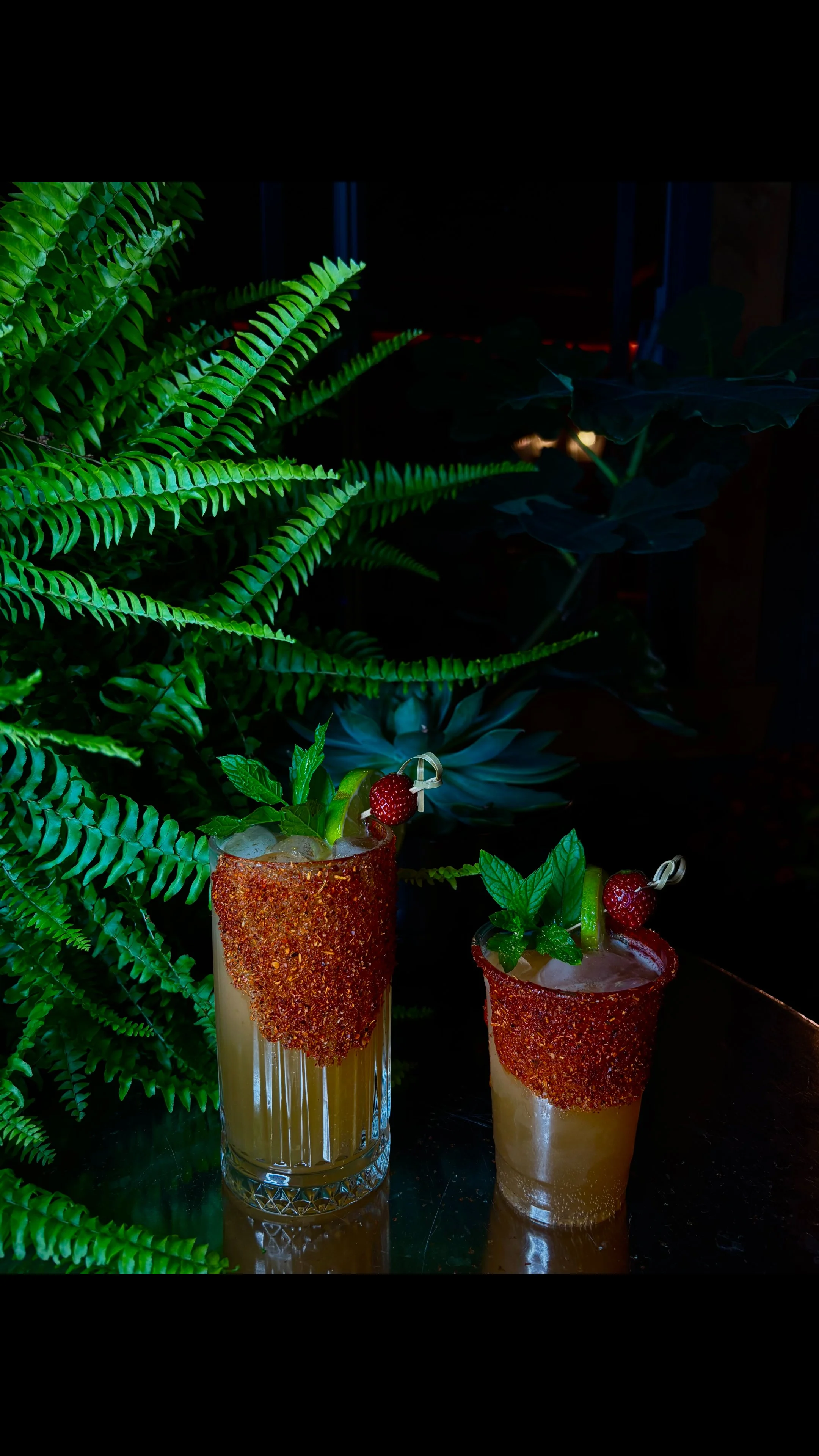 Two cocktails with tajin rim, garnished with mint, lime, and strawberry, placed on a reflective surface surrounded by green plants.