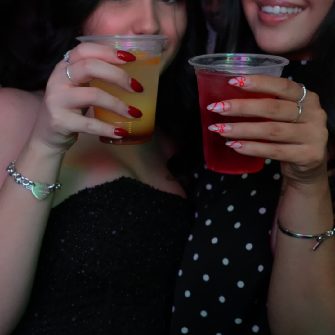 Two women holding drinks in plastic cups, one with a yellow-colored beverage and the other with a red-colored beverage, smiling and wearing holiday-themed nail art and jewelry.