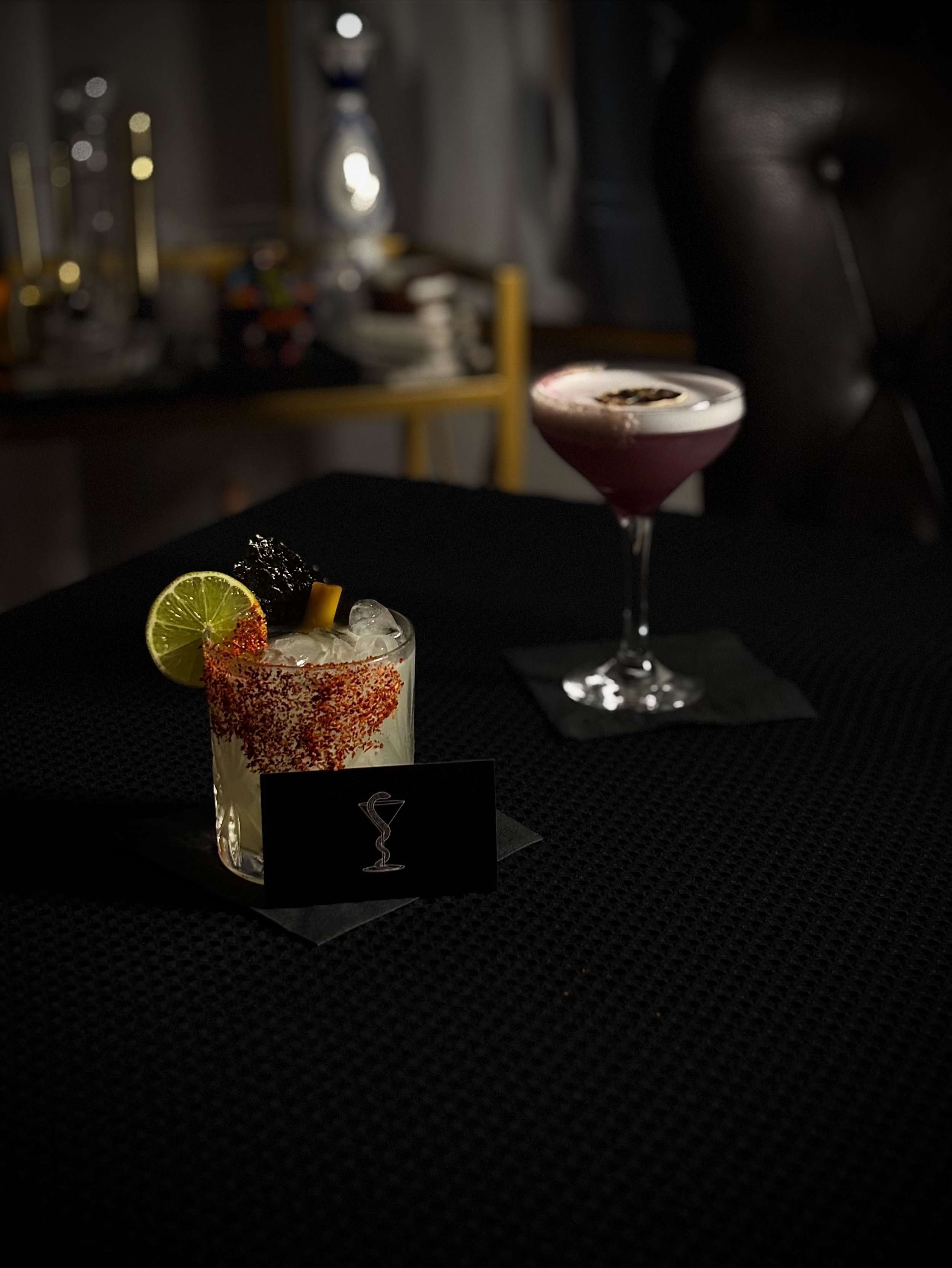 Two cocktails on a black textured table with a dark background and blurred bar equipment. One cocktail is a short glass with a lime wedge, chili powder rim, and ice, and the other is a pink martini in a stemmed glass.