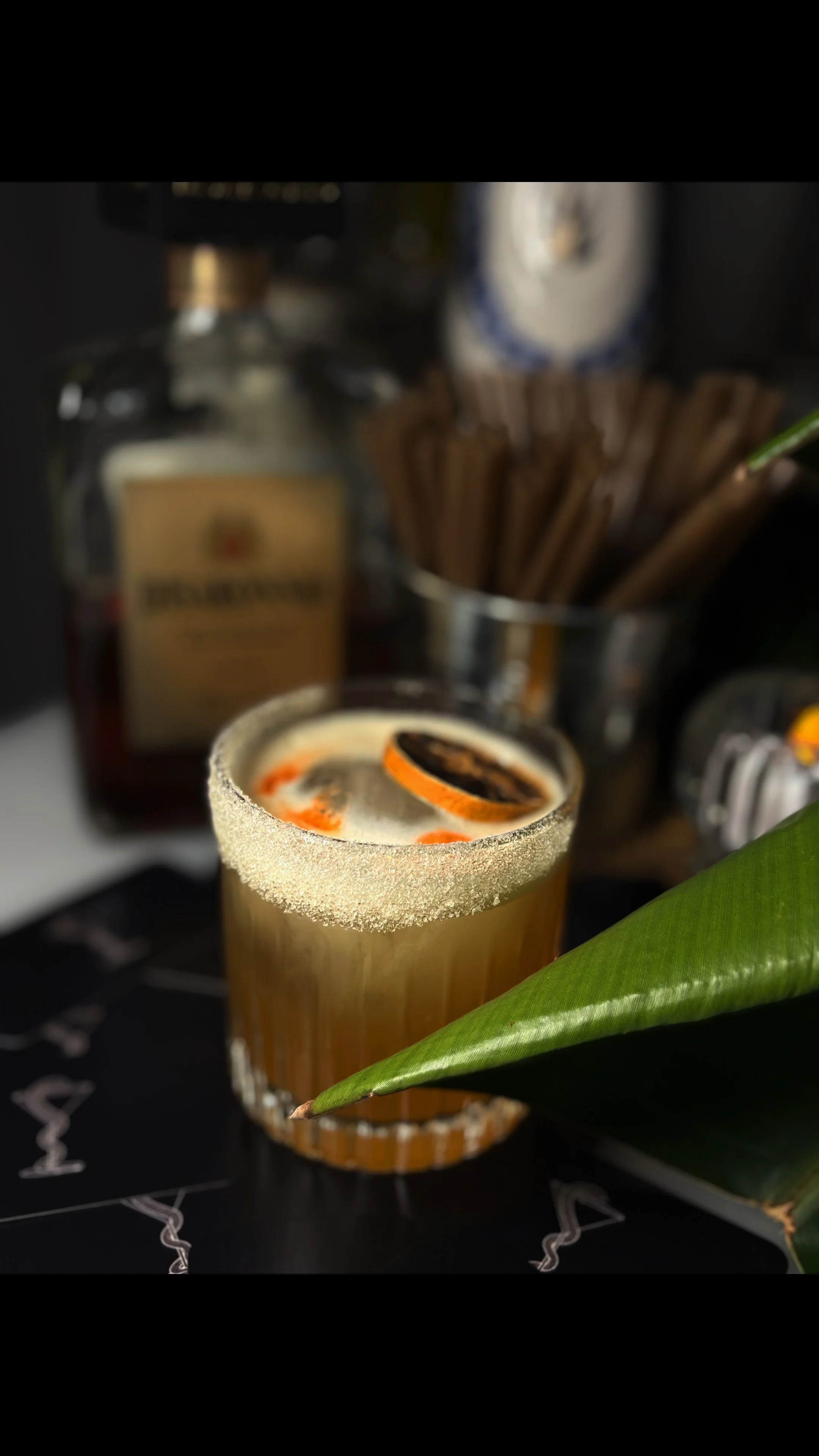 An amaretto sour with a sugared rim, garnished with a dried orange slice and hearts made from angostura bitters, set on a black surface with a large green leaf in the foreground and a blurred bar background.