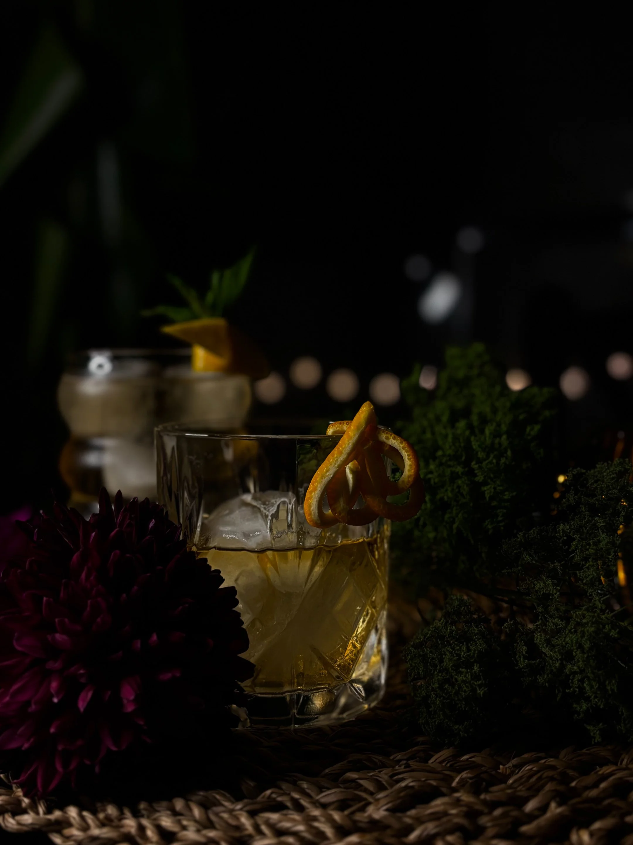 A glass of whiskey with ice and orange peel garnish, with a second drink in the background, decorated with lemon and orange wedges, surrounded by dark flowers and foliage on a woven surface.