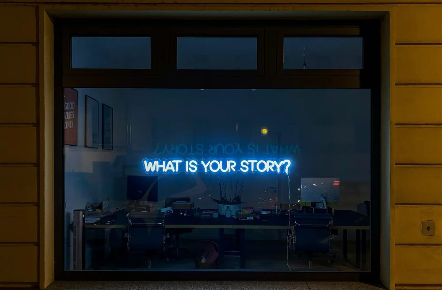 A window showing a room with a neon sign that reads 'WHAT IS YOUR STORY?' in blue, with some chairs and tables inside.