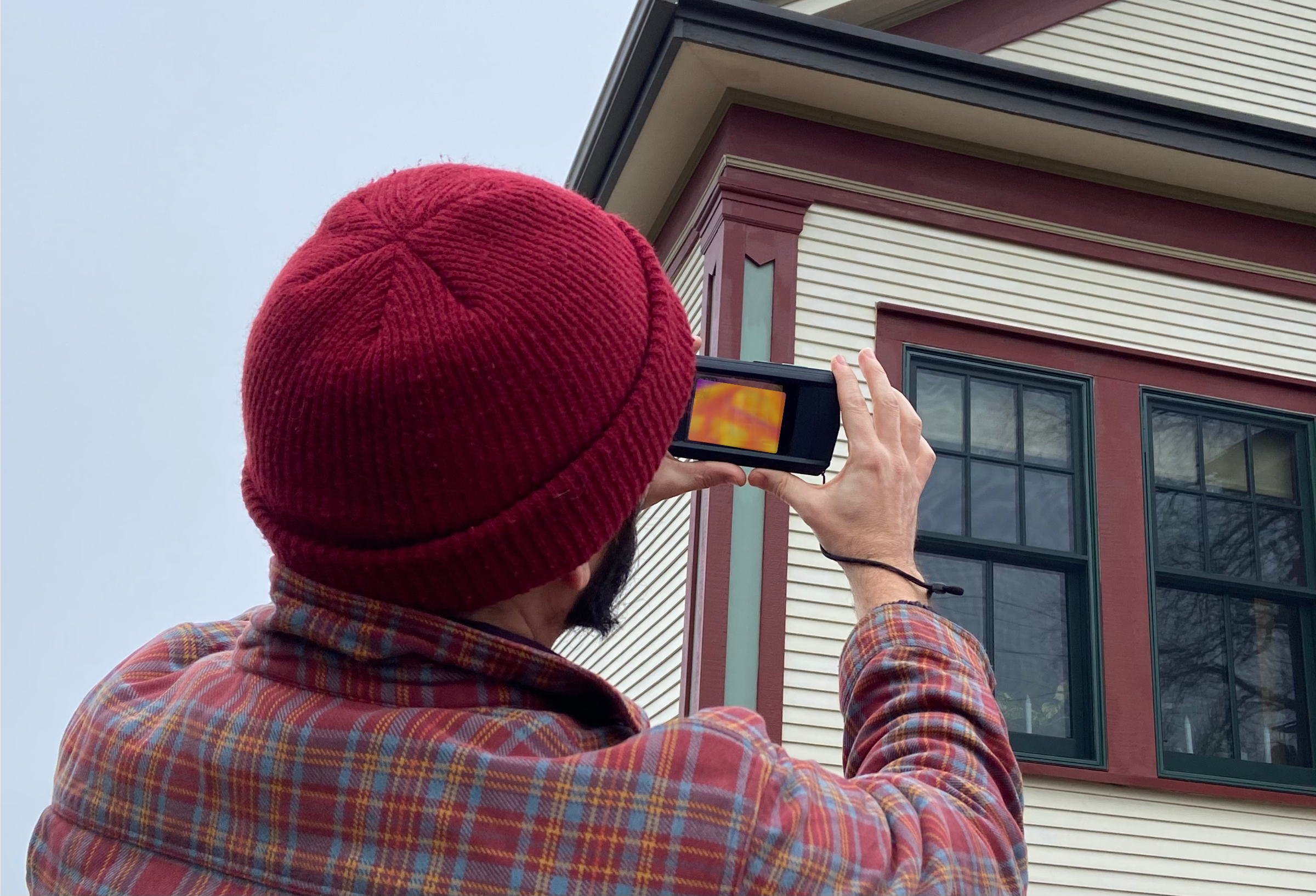 A person wearing a red knit beanie and a plaid shirt takes a thermal image of a house's window with a handheld thermal camera at a Tazewell County area historical home.