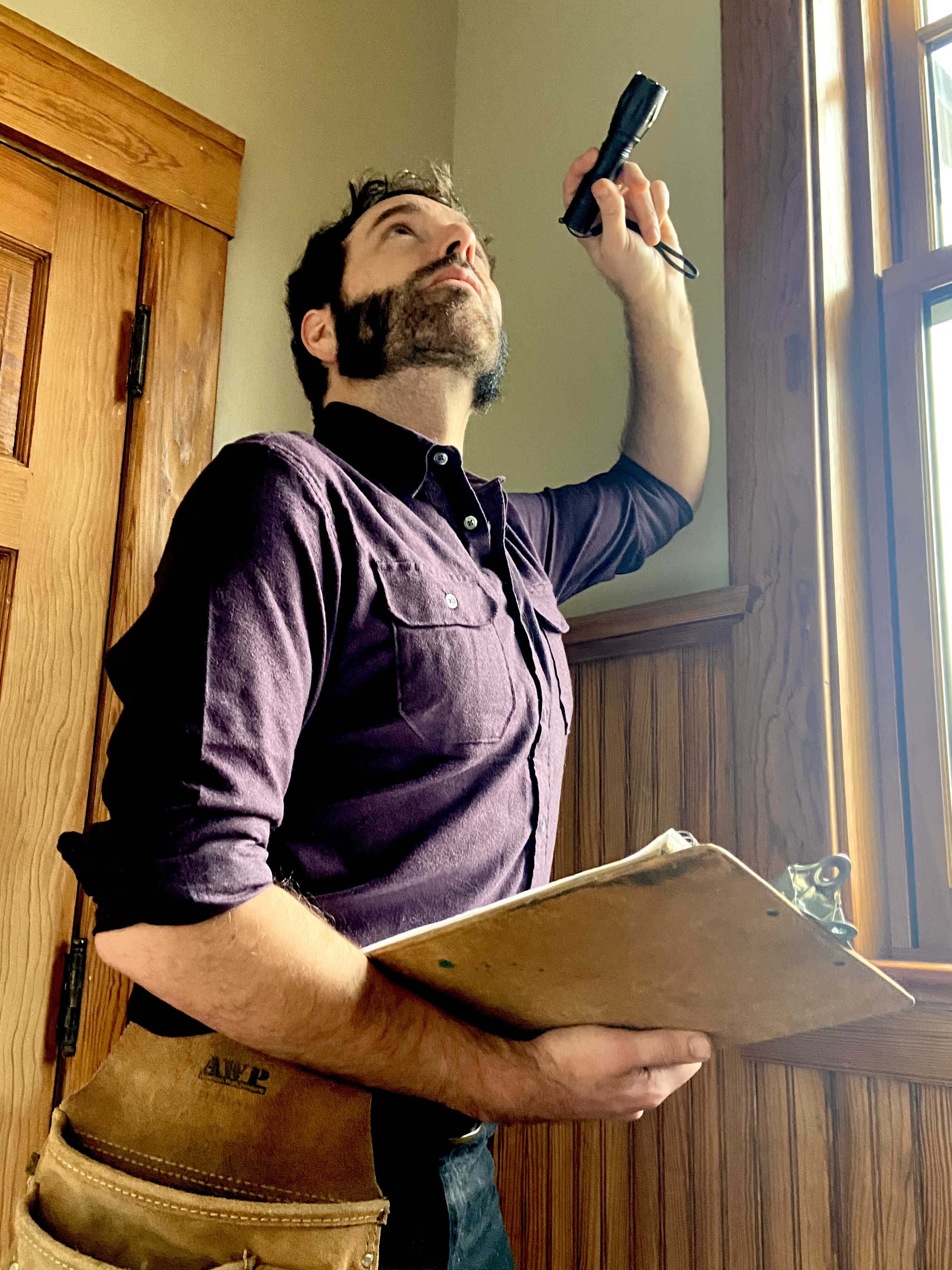 A man with a beard wearing a button-up shirt holding a clipboard, standing near a window, using a flashlight. The background behind him is a historical wooden door, window, and trim in a Peoria historical home.