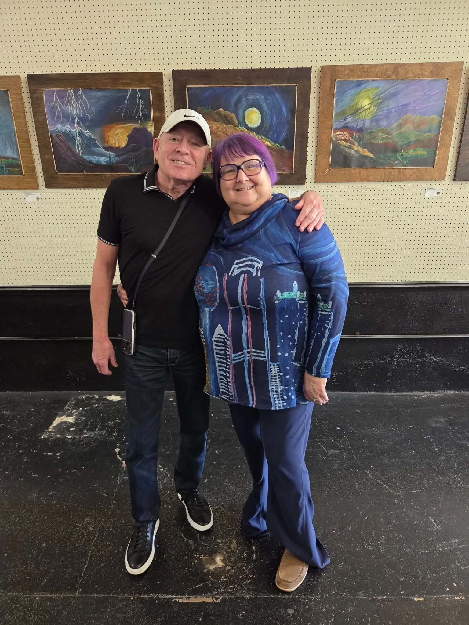 A man and woman smiling and hugging in an art gallery with colorful landscape paintings on the wall behind them.