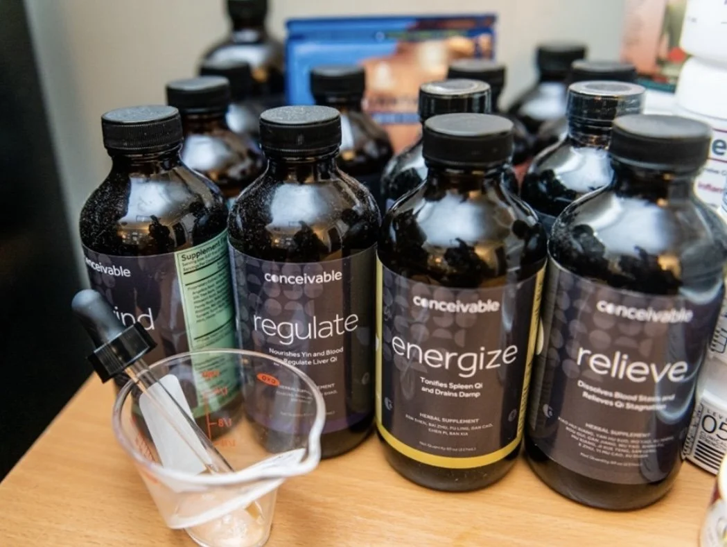 A collection of black supplement bottles labeled "conceivable" with word themes like "regulate," "energize," and "relieve" on a wooden surface, alongside a small glass measuring cup with a dropper in it.