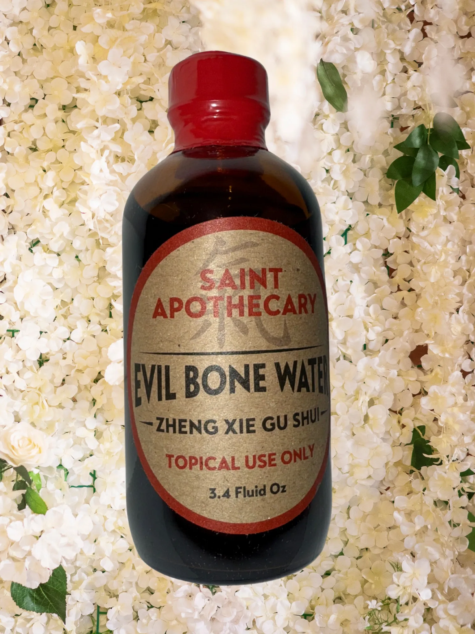 A brown bottle labeled 'Saint Apothecary Evil Bone Water' with red cap, placed against a background of white flowers and green leaves.