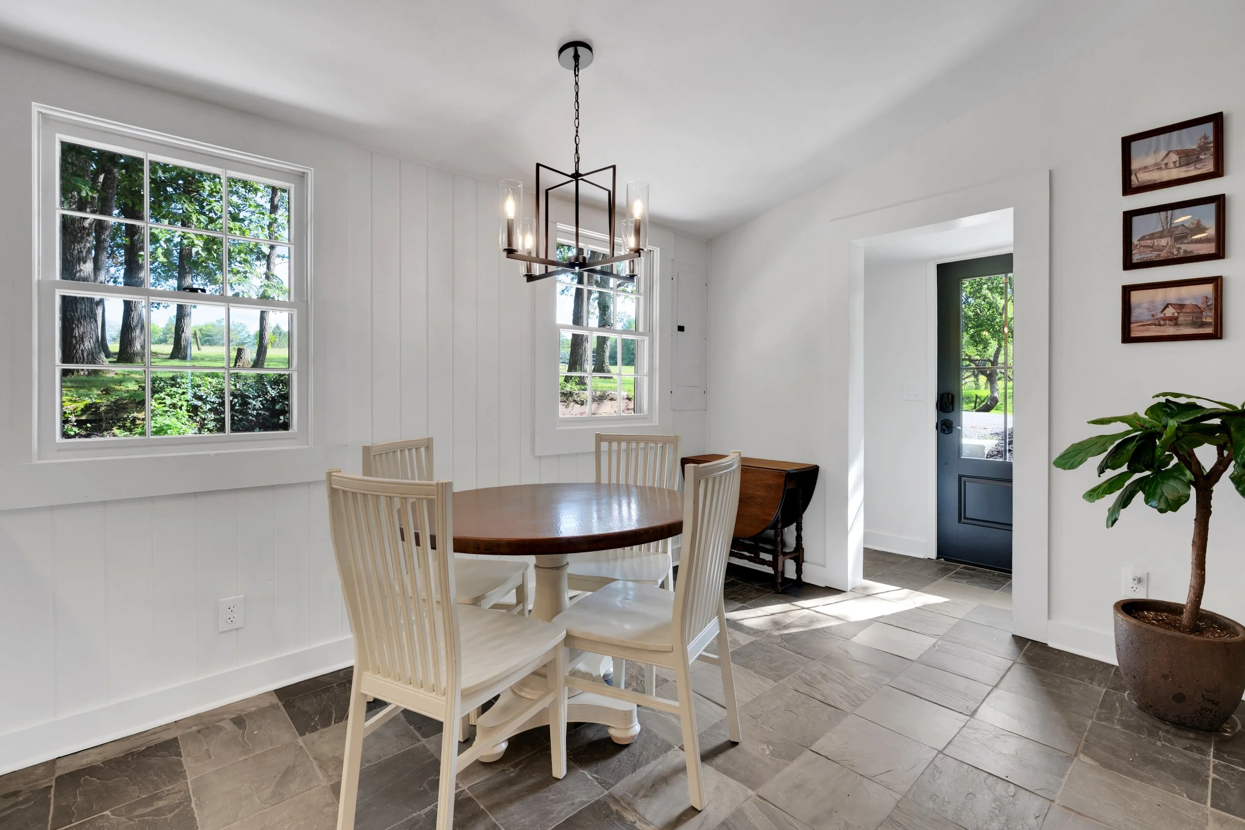 Dining room with round wooden table and beige chairs, white walls, large windows showing greenery outside, black door with glass panel, chandelier hanging above the table, potted plant in corner, framed pictures on wall.