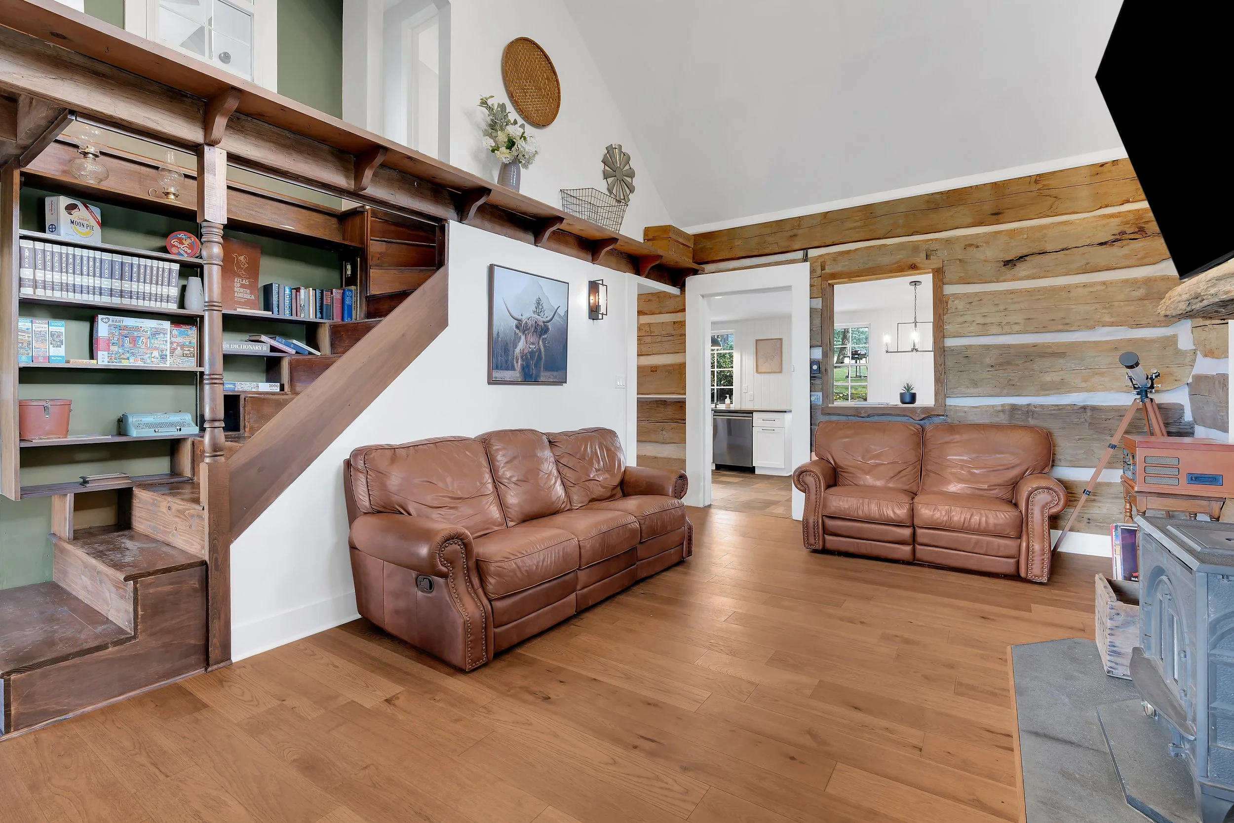 Living room with two brown leather sofas, wood flooring, a wood and stone fireplace, and a rustic wood wall. A staircase with built-in shelves is on the left side, and there's a botanical painting and wall decor.