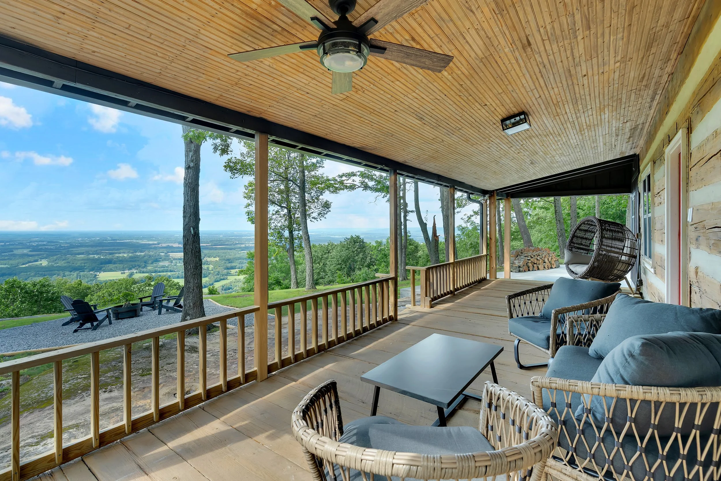 Covered porch with wooden ceiling and railing, outdoor furniture including sofas, chairs, and a hanging pod chair, overlooking a landscape with trees, hills, and a valley.