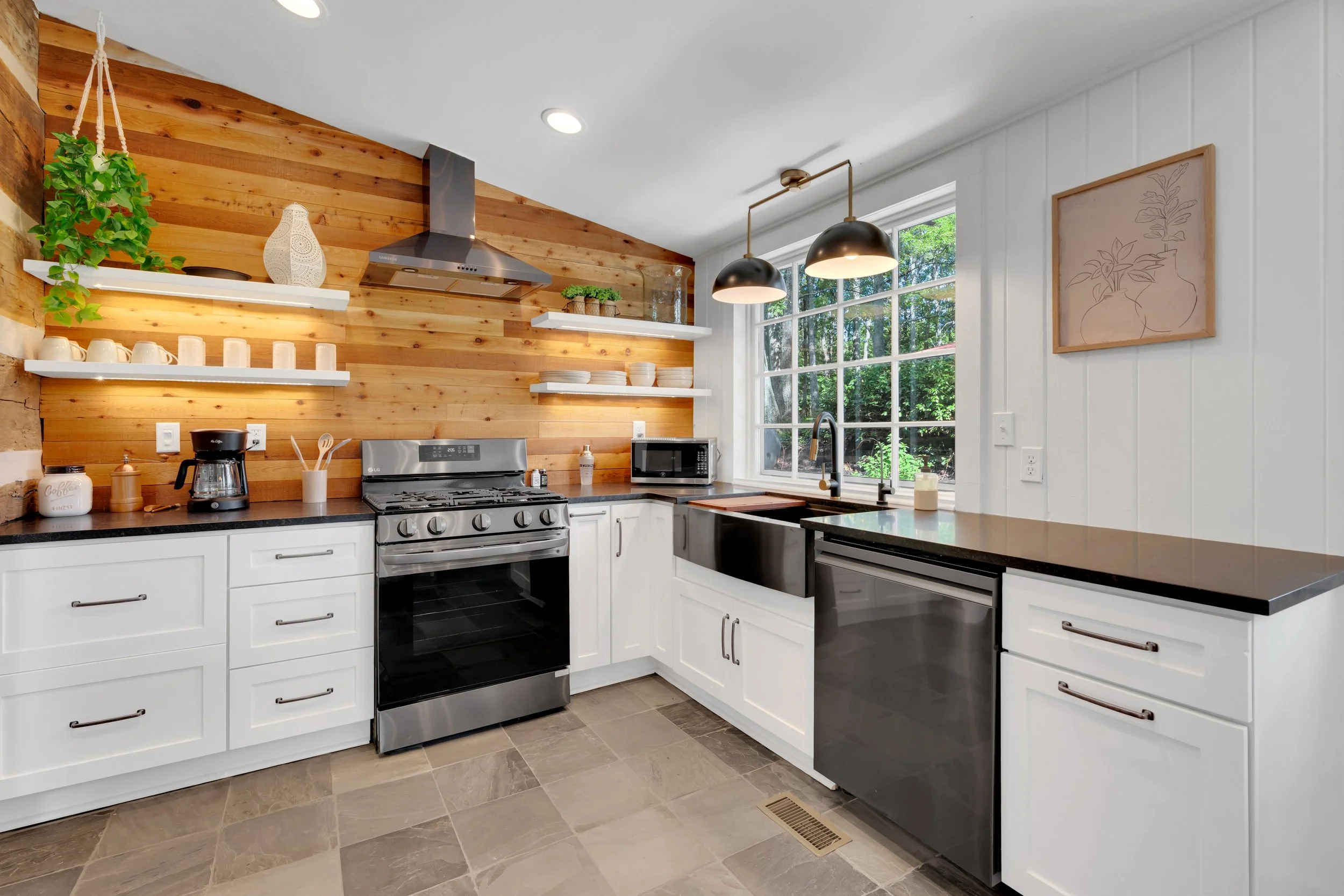 Modern kitchen with white cabinets, black countertops, stainless steel appliances, wooden accent wall, open shelves, large window, and pendant lighting.