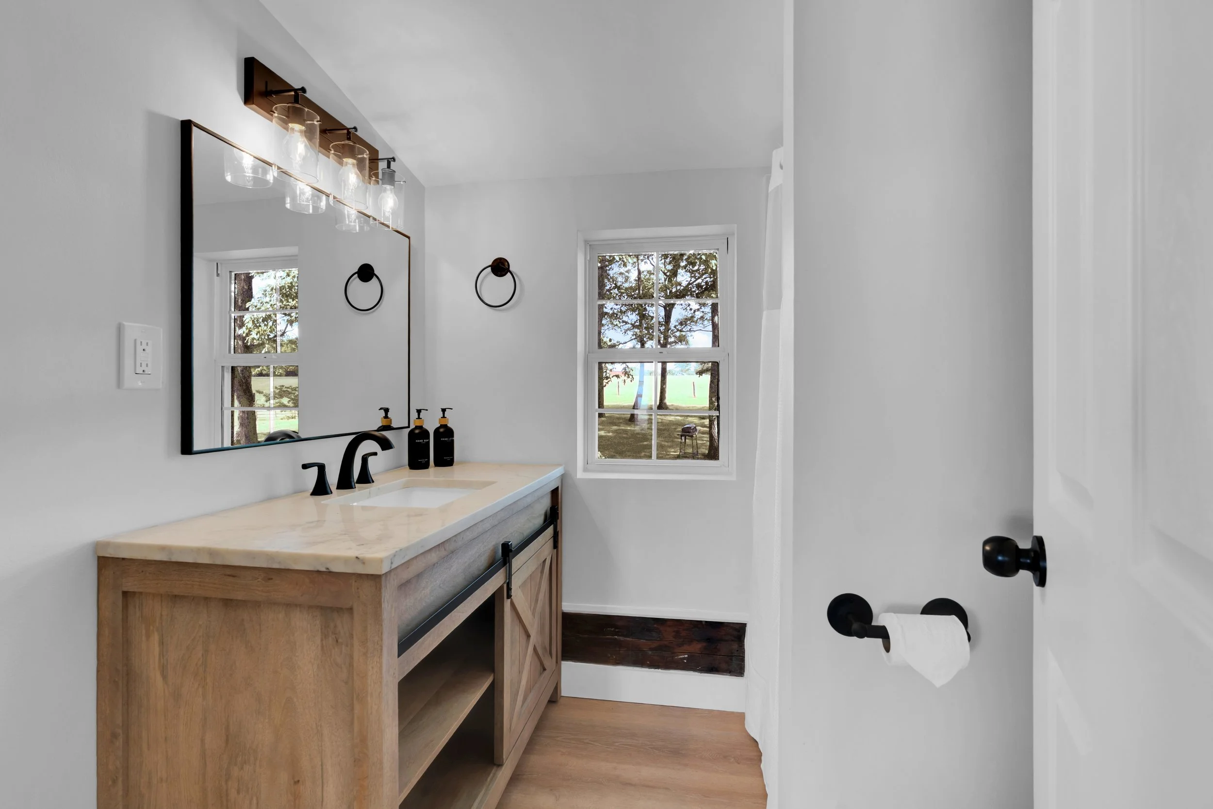 Modern bathroom with wooden vanity, black faucet, large mirror, two soap dispensers, window showing trees outside, white walls, black towel ring, and toilet paper holder.