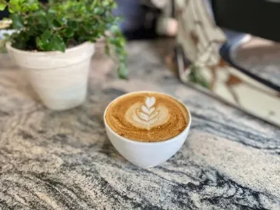 A cup of coffee with latte art on a marble table, with a potted plant and a person in the background.