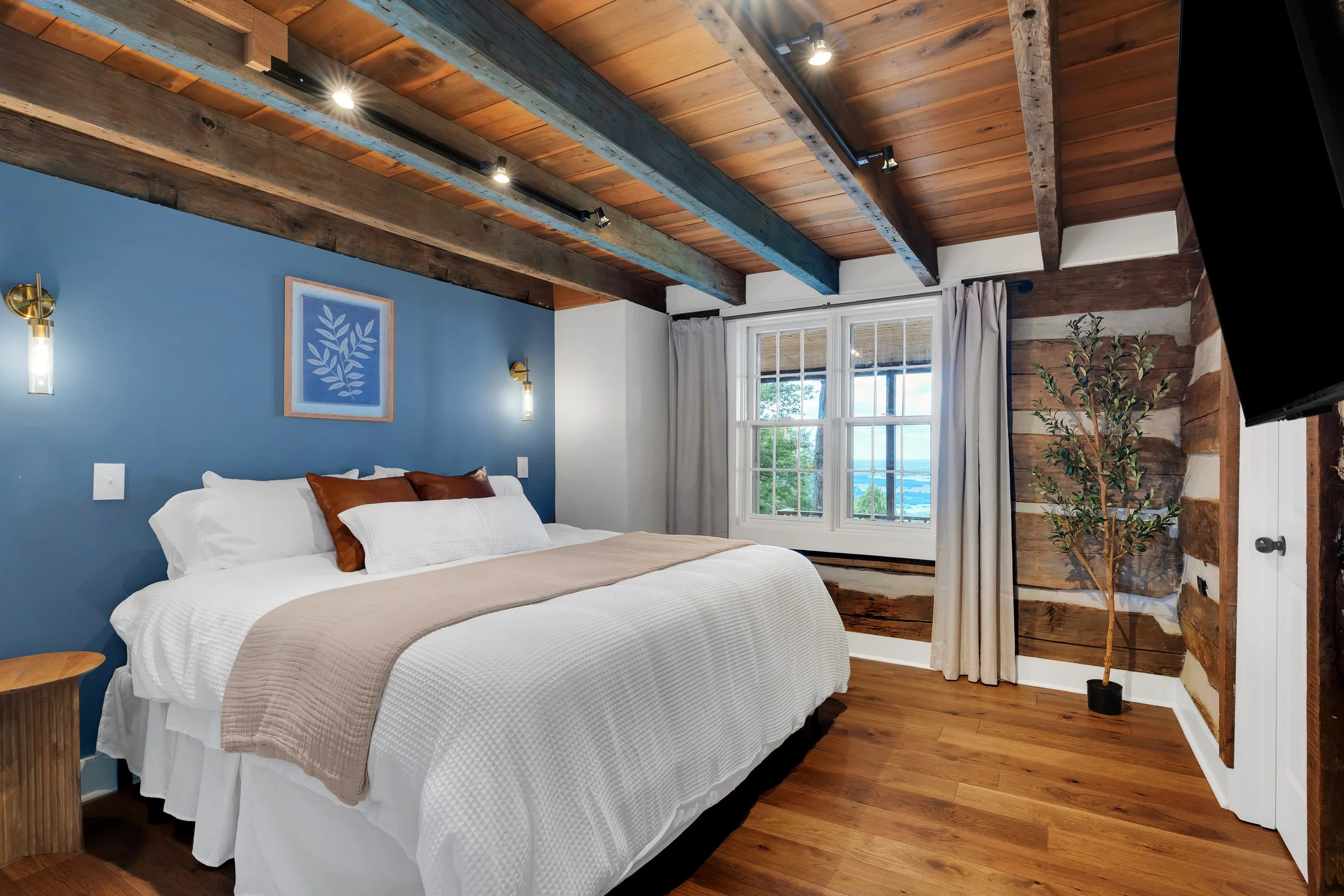 A cozy bedroom with wooden ceiling beams, blue accent wall with framed art, large window with white curtains, and a bed with white bedding and brown pillows.