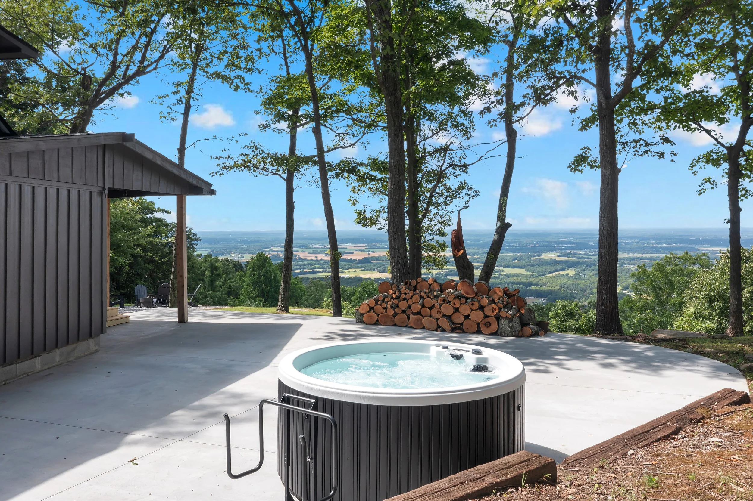 Outdoor hot tub on a concrete patio with a view of trees, a distant landscape, and blue sky, surrounded by tall trees and stacked firewood. Gorgeous views