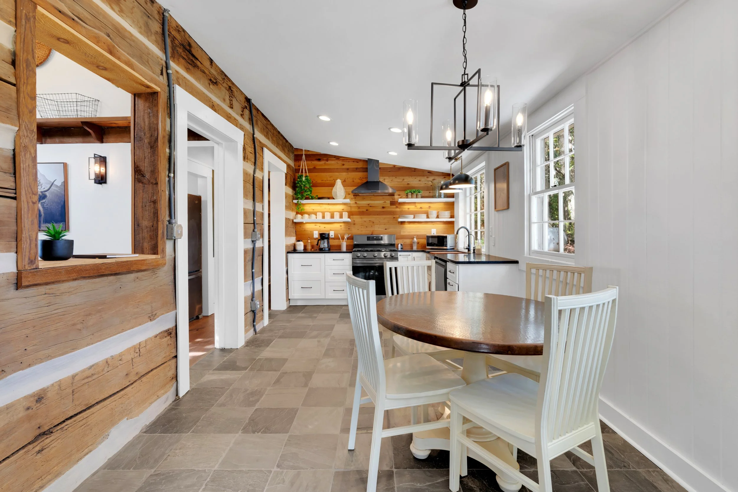 A bright kitchen with white cabinets, black countertops, and wooden accents. There is a round wooden dining table with white chairs, large windows allowing natural light, and a black chandelier hanging above the table.