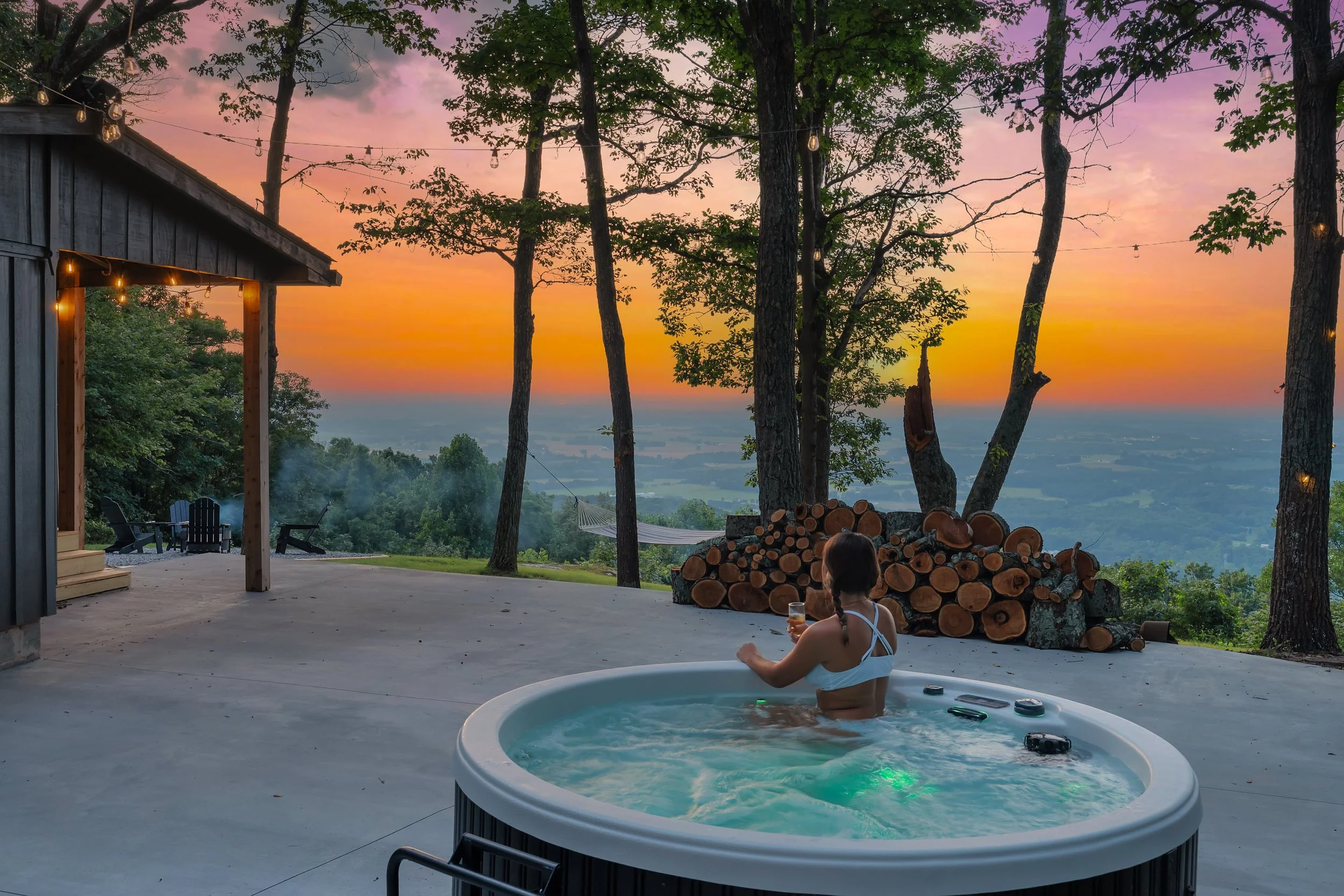 A woman relaxing in a hot tub on a patio during sunset, with trees, a woodpile, and a scenic view of the landscape in the background.