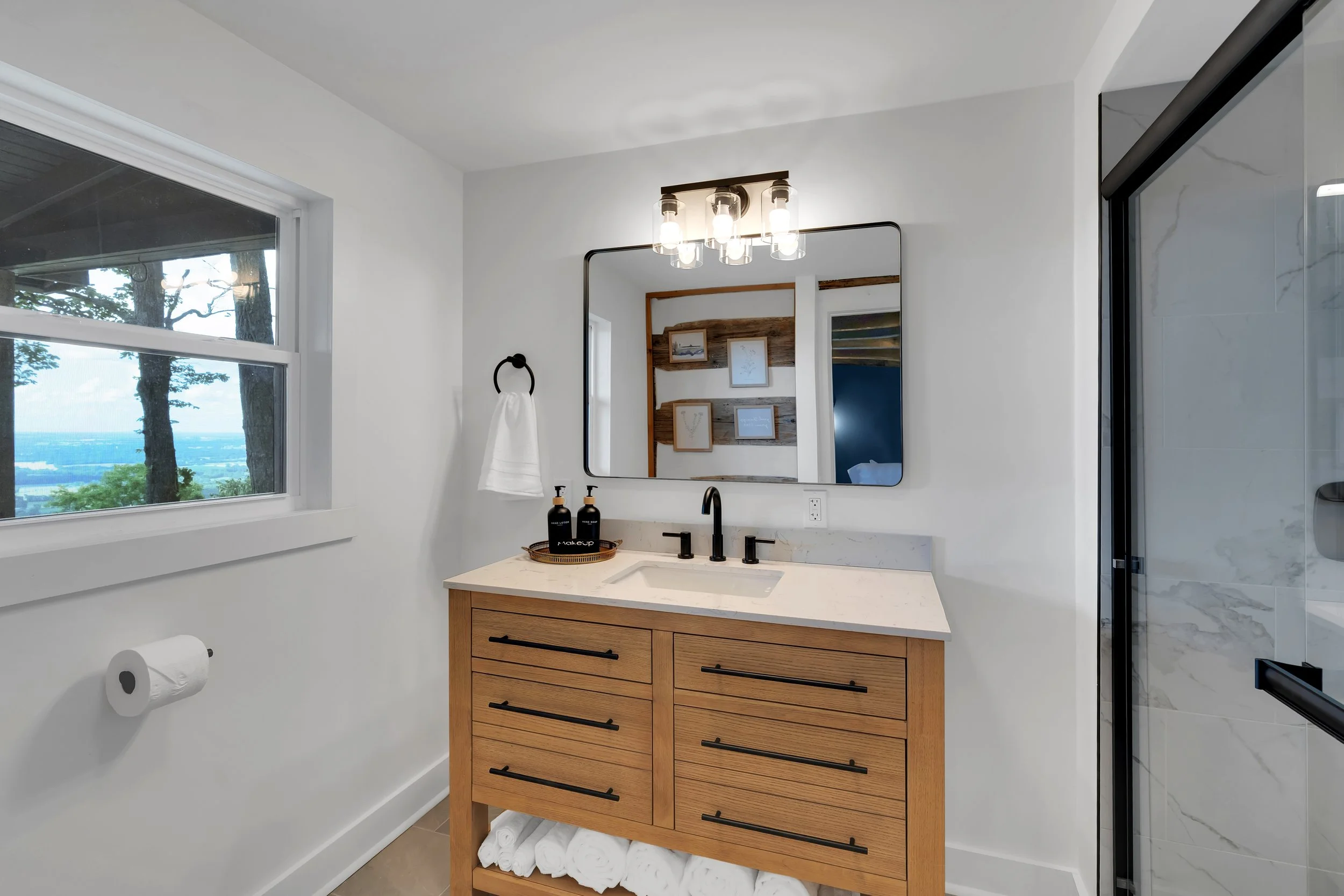 Modern bathroom with a wooden vanity, marble countertop, black faucet, large mirror, window showing trees, wall art, and a shower with glass door.