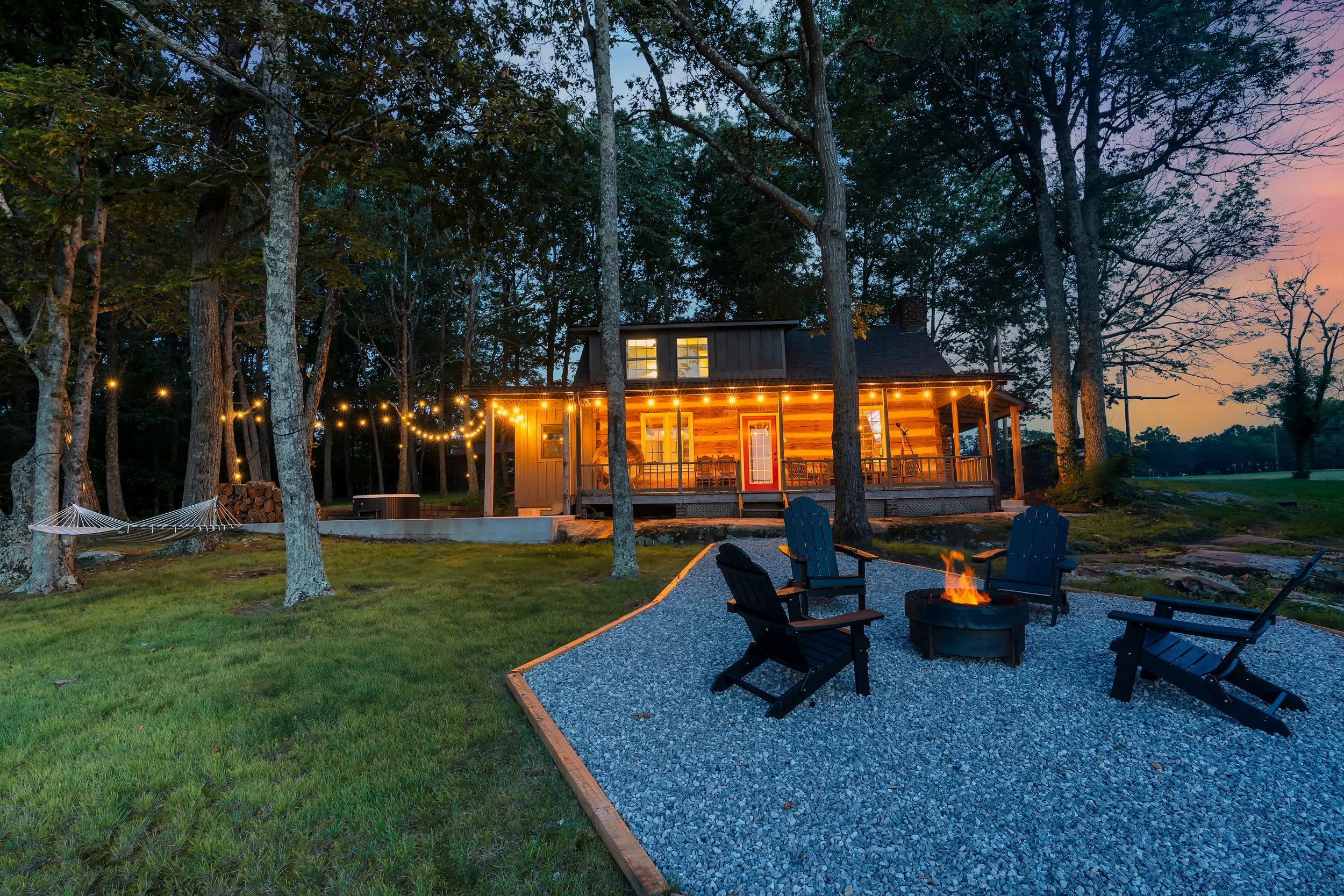 A cozy backyard with a fire pit surrounded by four Adirondack chairs, a gravel area, a house with string lights and a porch, trees, a hammock, and a sunset sky.